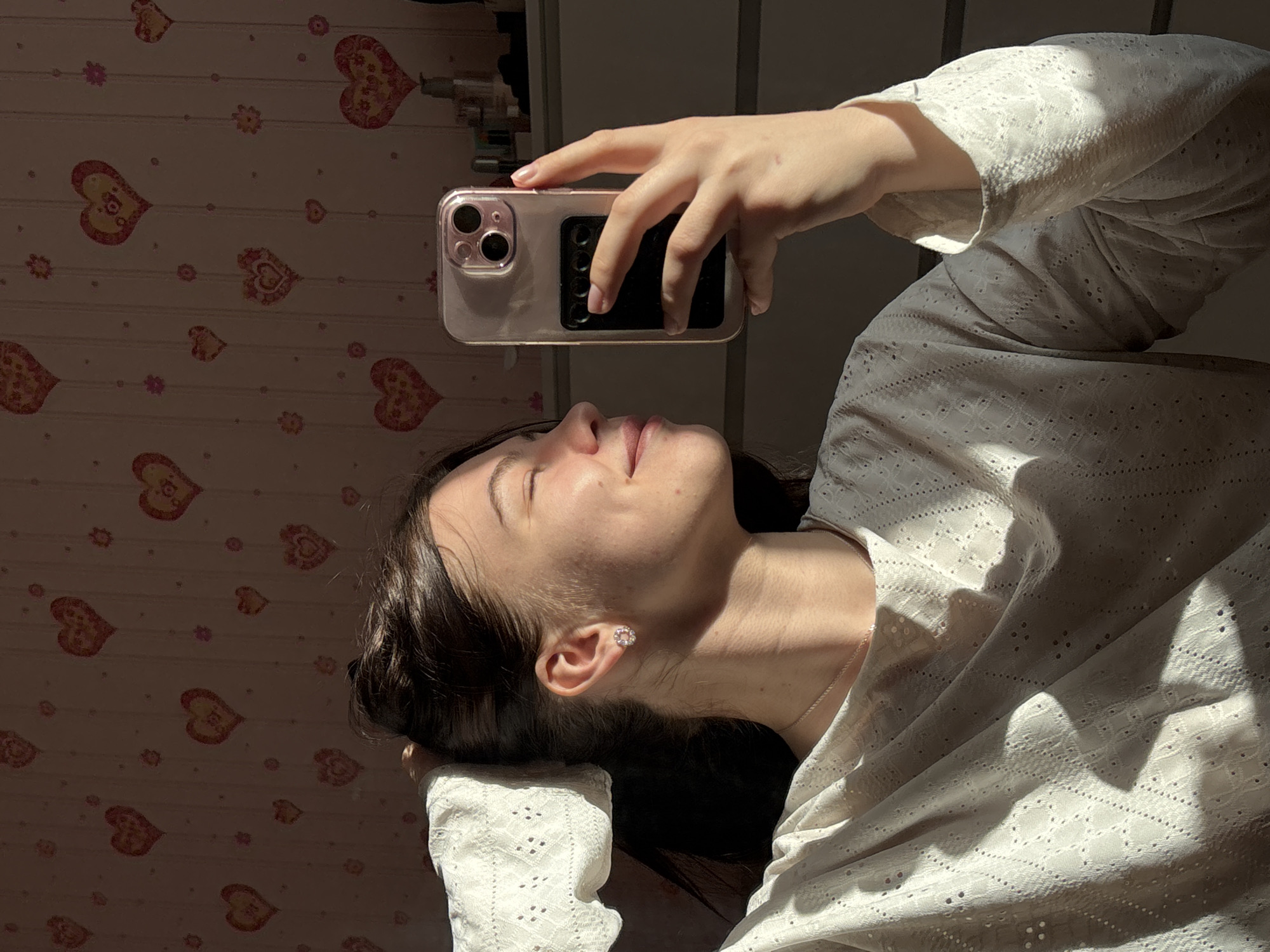 A young woman is lying on her side in Perm, Russia, holding a rose gold iPhone with a black grip on the back above her face. She is wearing a white, lacy blouse and a small earring in her left ear. Her dark hair is partially visible behind her head. The background is a light-colored wallpaper with a pattern of hearts and small flowers. Bright sunlight is cast across the scene, creating strong shadows on the woman and the wallpaper. The top of a cabinet or shelf can be partially seen at the top of the image.
