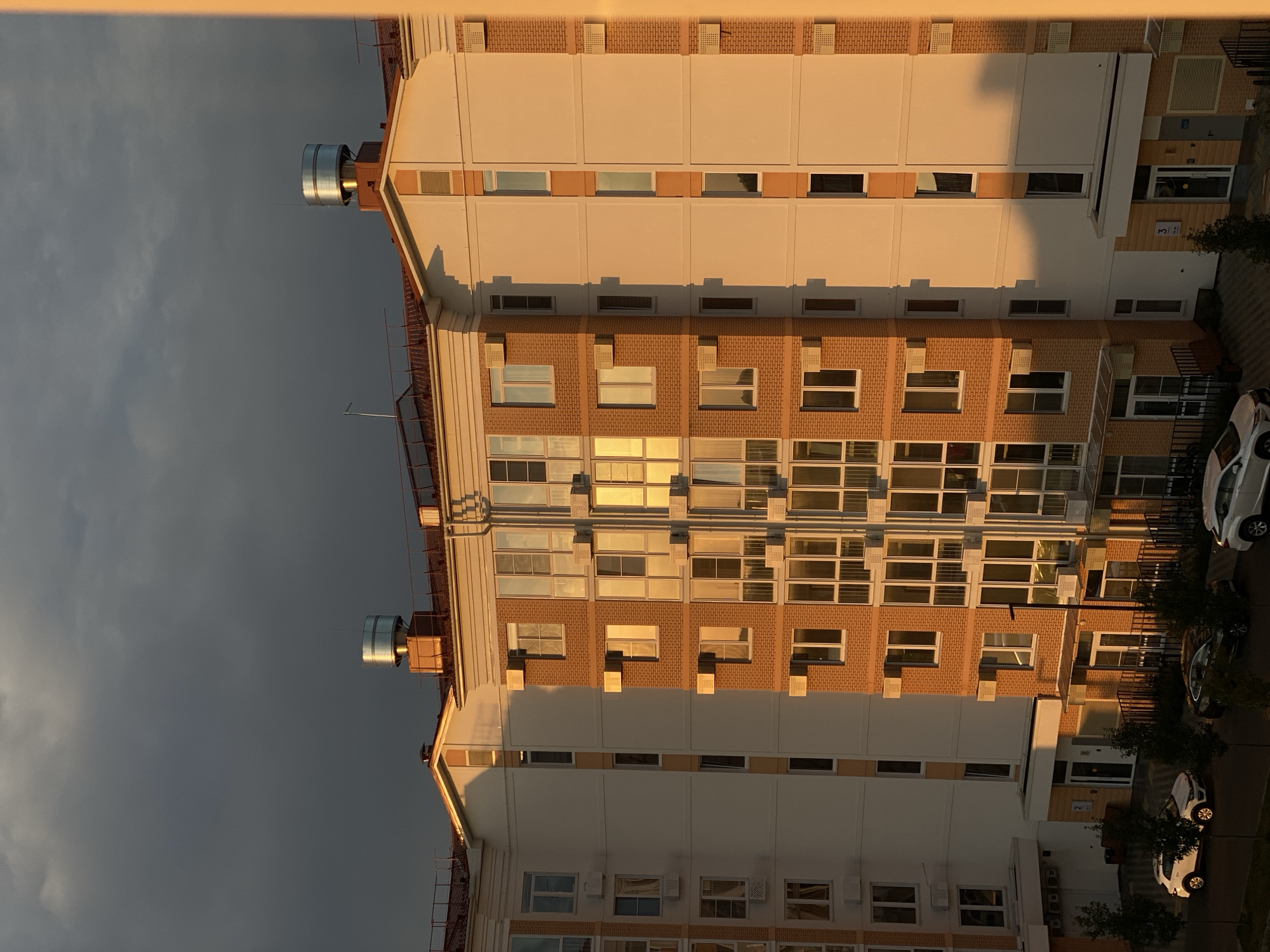 A multi-story residential building with a predominantly white and orange brick facade is viewed from an elevated position at sunset. The building features numerous windows, some reflecting the golden light of the setting sun, and air conditioning units mounted on the exterior. The roofline is adorned with what appear to be ventilation systems or exhaust pipes. In the foreground, parked cars, including a white SUV and a dark sedan, are visible on a paved area adjacent to the building. The sky above is partially overcast with dark clouds, contrasting with the warm lighting on the building. The scene suggests an urban or suburban setting in Dinskaya, Russia, during the evening.