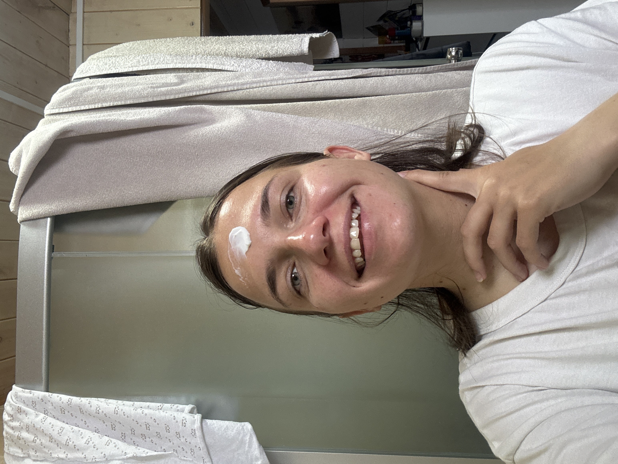 A young woman with dark hair is captured in a close-up, smiling broadly at the camera, conveying a sense of happiness and contentment. A distinct dollop of white cream or product is visible on her forehead, indicating she is in the process of a skincare routine. Her skin appears radiant and slightly dewy. She is wearing a simple white t-shirt, and her right hand is casually resting near her neck or shoulder.
The setting appears to be an indoor space, likely a bathroom or a personal care area, located in Раздолье, Russia. In the upper background, several light-colored, possibly beige or white, towels are neatly folded and stacked on what looks like a shelf or the top of a shower enclosure. Behind the woman and to her right, a large, light-grey, smooth surface, which could be a shower wall or a mirror, occupies a significant portion of the background. To her left, wooden paneling lines a wall, adding a rustic touch to the environment. In the lower left foreground, a white patterned cloth or towel is partially visible. There is no discernible text within the image.