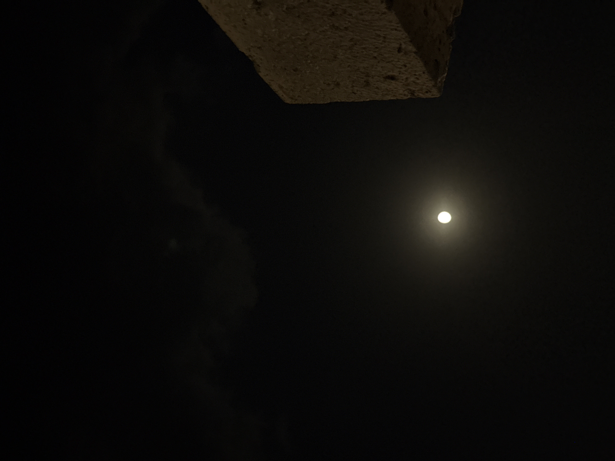 A full moon is visible in the dark night sky, positioned slightly left of center. A portion of a rough, light-colored structure juts into the frame from the upper right corner. The scene is dark, with some faint cloud cover visible in the upper portion of the sky. There are no people or animals present. The image appears to be taken in an outdoor urban or semi-urban environment.