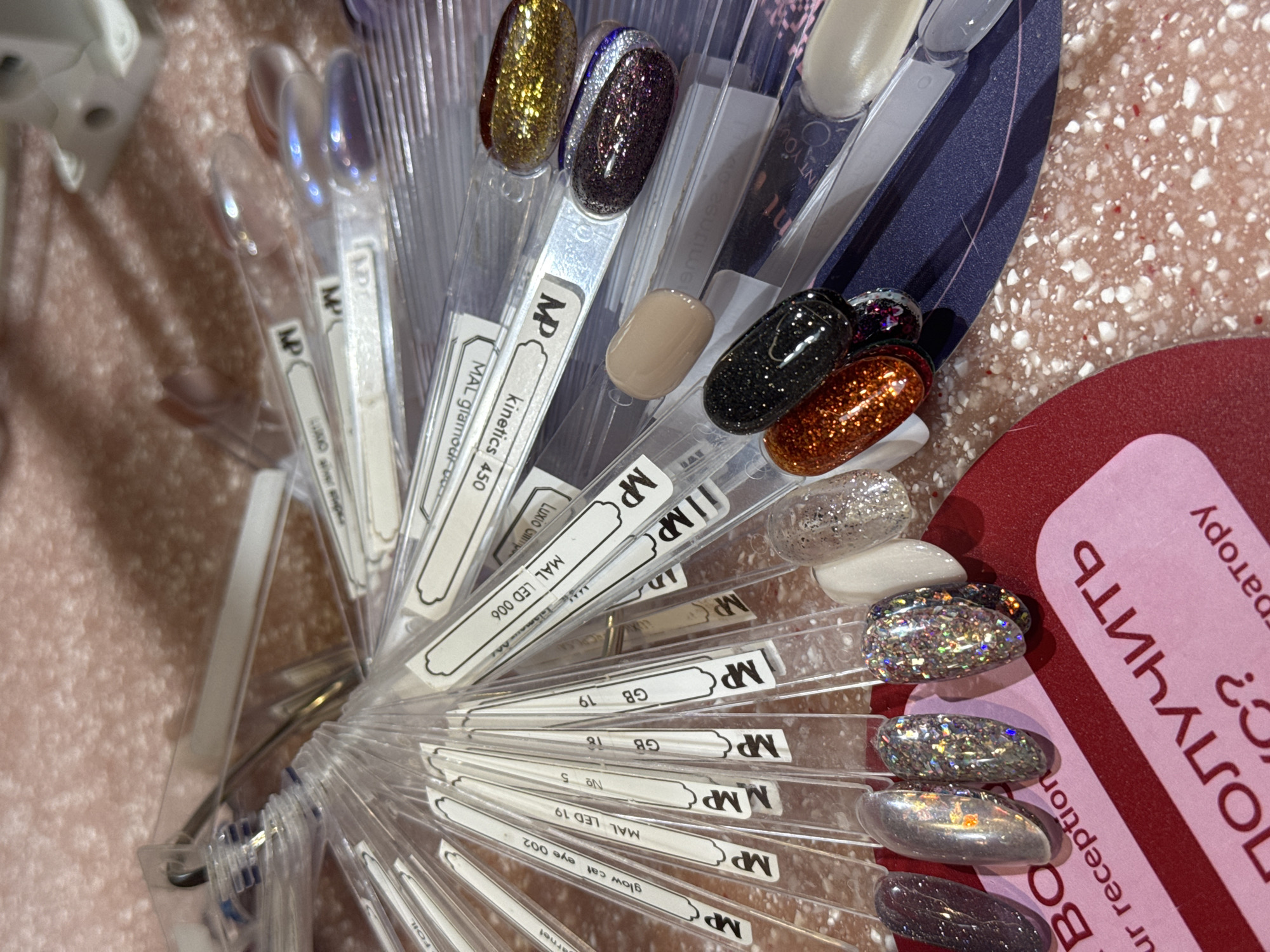 The image shows a nail polish display arranged in a semi-circular fan. The polishes vary in color and texture, including glitter, metallic, and solid finishes. Each polish is attached to a clear plastic stick with a small label at the base. The labels are marked with the letters "MP," and some include additional identifiers such as "MAL LED 006," "Luxio Glitter," "kinetics 450," and "MAL glamour-cool." A red and pink sign with text in a foreign language is partially visible on the right side of the image. The surface beneath the display is a light-colored speckled material. It is not possible to infer a location in the city of Dubai, United Arab Emirates, from this image.