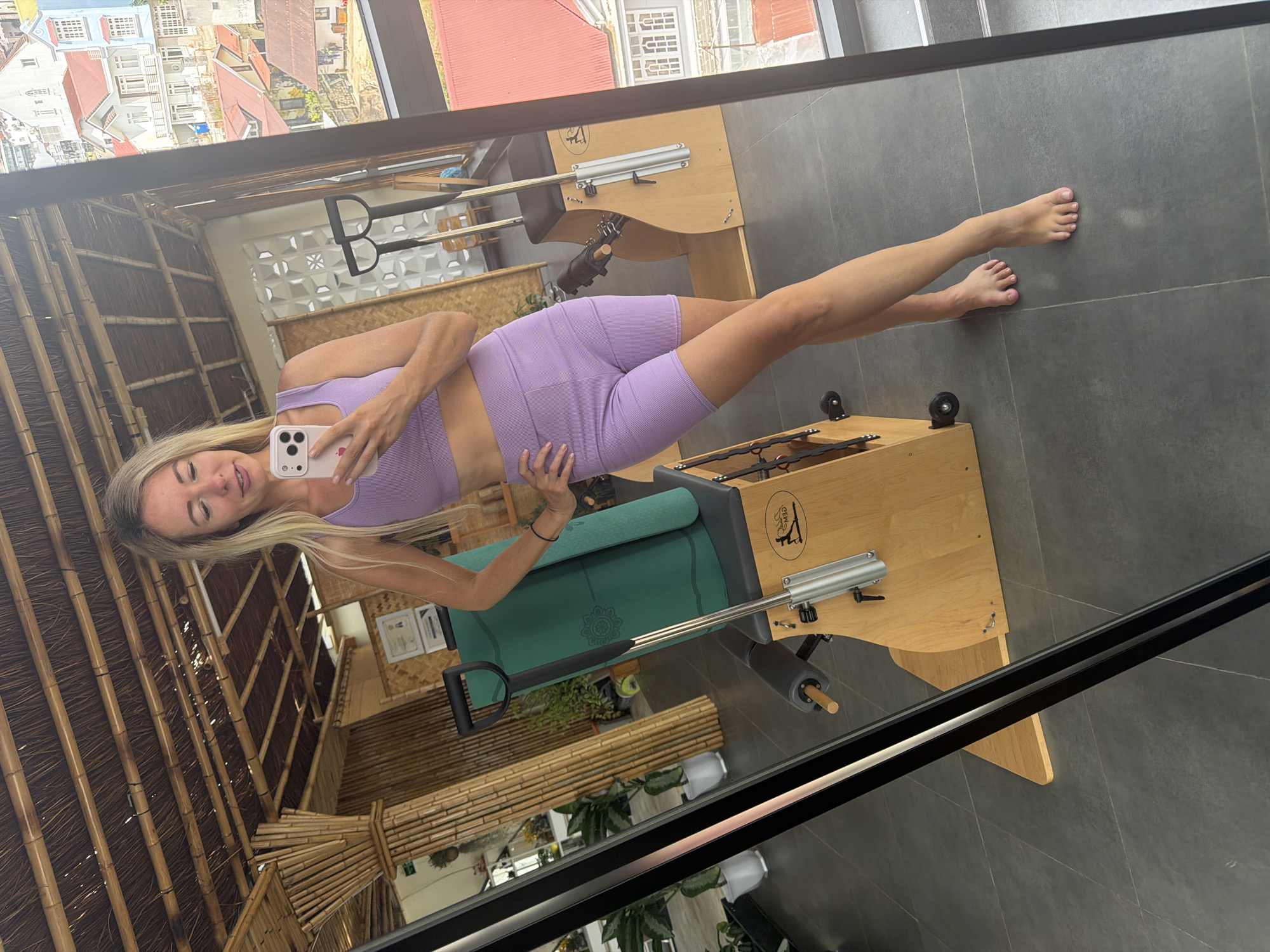A woman with blonde hair and a purple athletic outfit is taking a mirror selfie in a room with a bamboo ceiling and walls. She is holding a smartphone in her right hand and has her left hand on her hip. In the background, there are pilates reformers and plants. The room appears to be a fitness studio located in Da Lat, Vietnam, with a view of colorful houses and buildings visible through a large window.