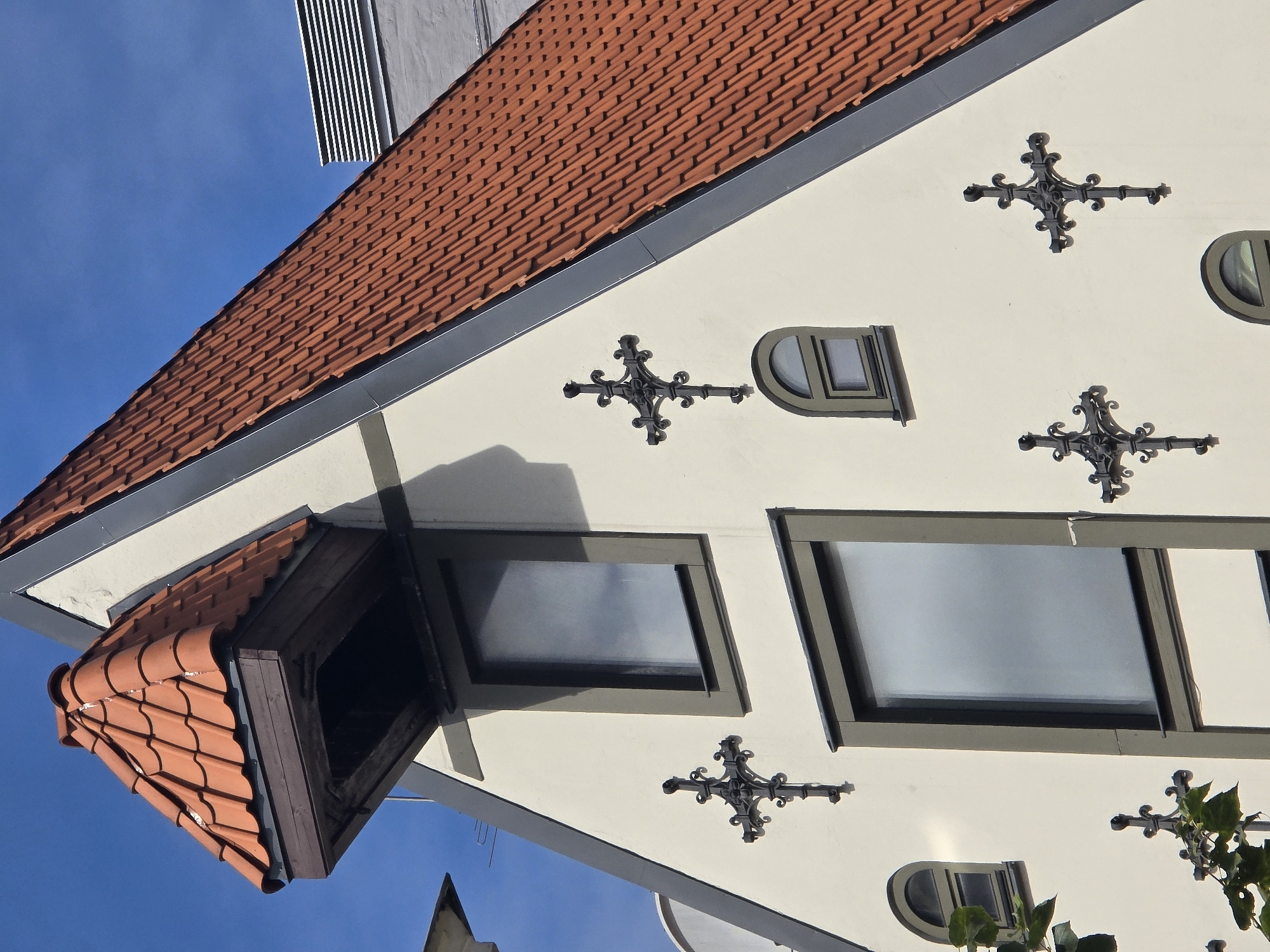 This image shows a close-up, eye-level view of the upper facade of a historic building in Riga, Latvia. The building features a prominent steeply pitched roof covered in red clay tiles, with a small dormer window jutting out. The facade is painted a light cream color and is adorned with several decorative wrought-iron crosses. There are at least three rectangular windows and two small arched windows visible. The sky is clear and blue. The architectural style suggests a European, possibly Gothic or Neo-Gothic influence, common in the Old Town of Riga.