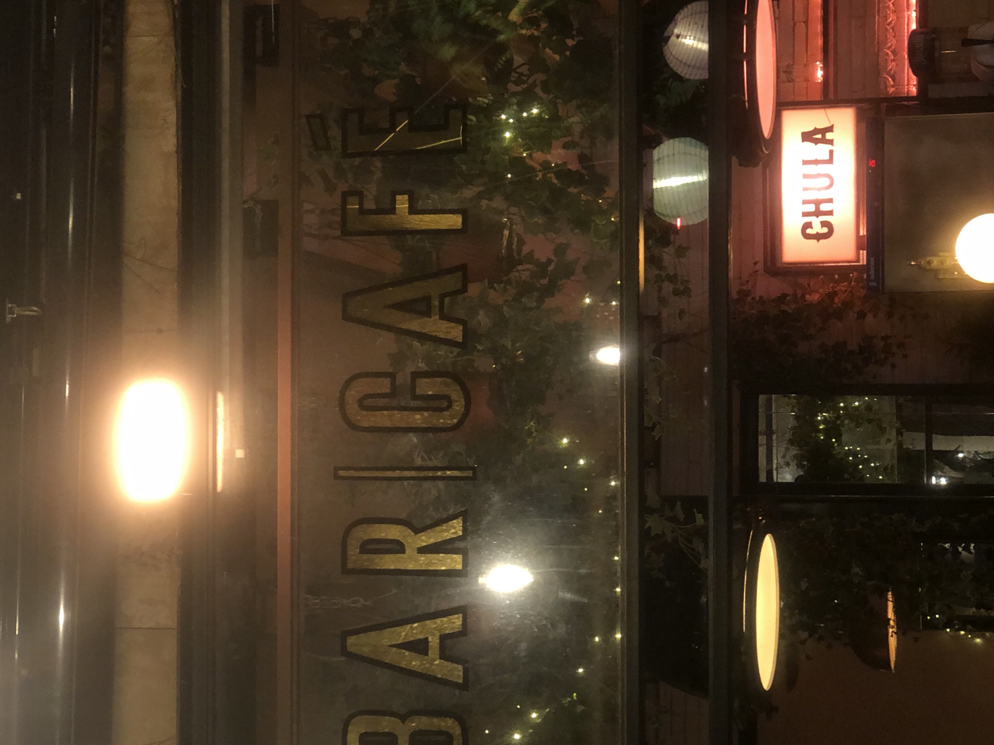 An exterior shot of a bar and cafe called "BAR | CAFE" at night. The sign is illuminated with gold lettering against a dark background, with decorative string lights visible behind it. Below the sign are some pendant lights and a smaller illuminated sign that reads "CHULA". The scene appears to be a street-level establishment.