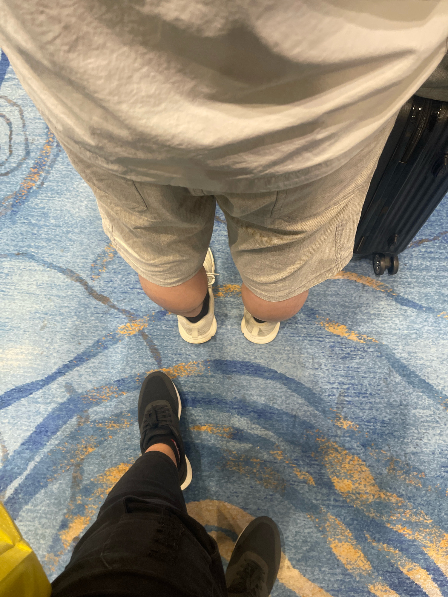 This is an aerial shot of two people standing on a blue carpet with yellow and white abstract patterns. The person closer to the camera is wearing black ripped jeans and black sneakers. Their left leg is bent at the knee, and their right foot is pointed forward. The second person, further away, is wearing light grey shorts, a light grey t-shirt, and white sneakers. They are standing next to a dark grey suitcase with black wheels. The environment appears to be an indoor public space, possibly an airport or hotel lobby, given the carpet design and the presence of luggage. The lighting suggests an indoor setting. No specific landmarks or elements of Singapore are visible in the image.