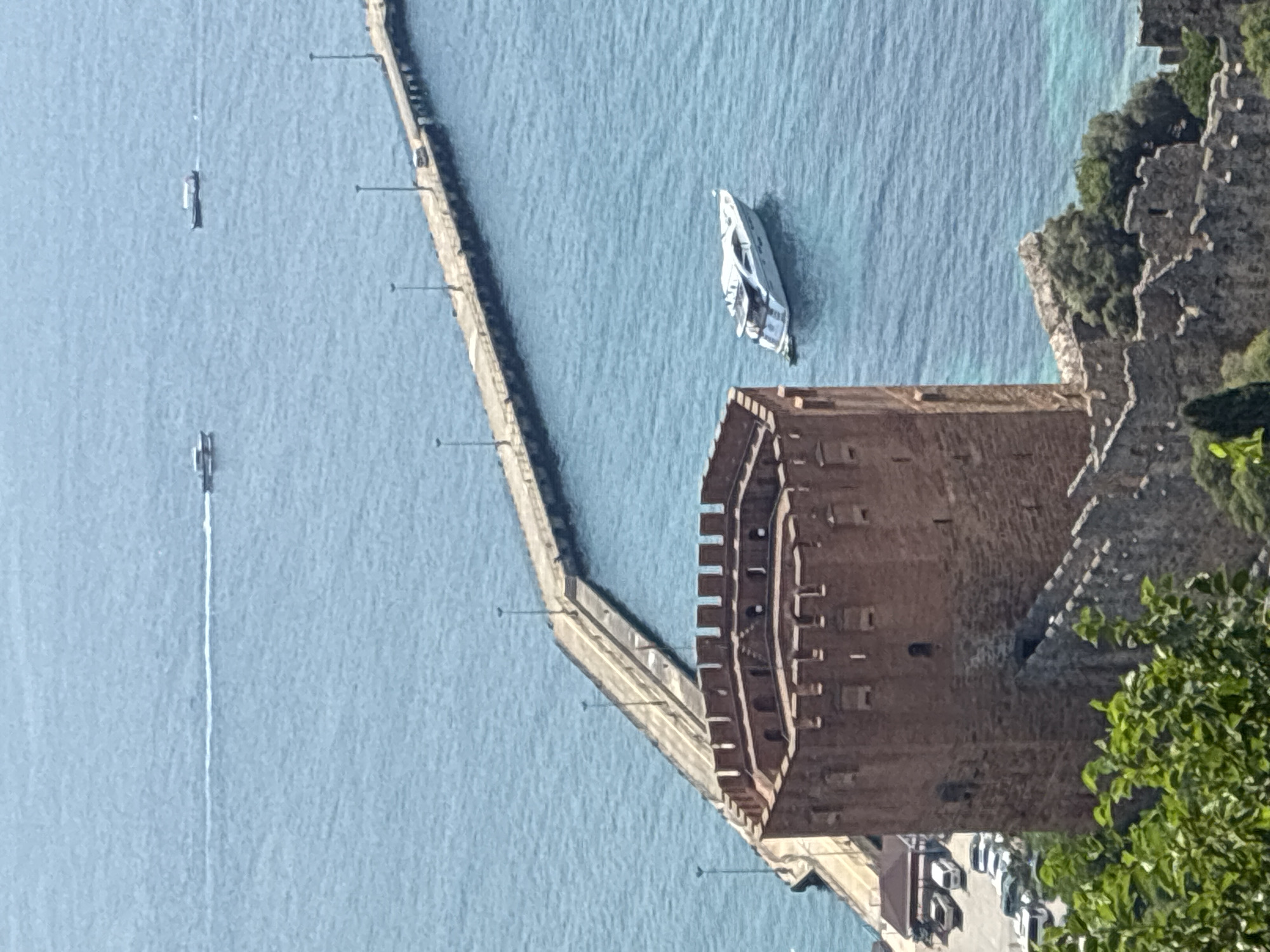 The image shows a scenic view of the Red Tower (Kızıl Kule) in Alanya, Turkey, overlooking the sea. The Red Tower, a prominent octagonal stone structure, stands in the foreground, its weathered red bricks contrasting with the clear blue water. A long pier extends into the sea from the left side of the image, lined with lampposts. Several boats are visible on the water: a modern white yacht is anchored near the pier, and two smaller boats are further out in the distance. The background reveals a hazy coastline and the vast expanse of the Mediterranean Sea. The scene appears to be captured on a bright, sunny day.
