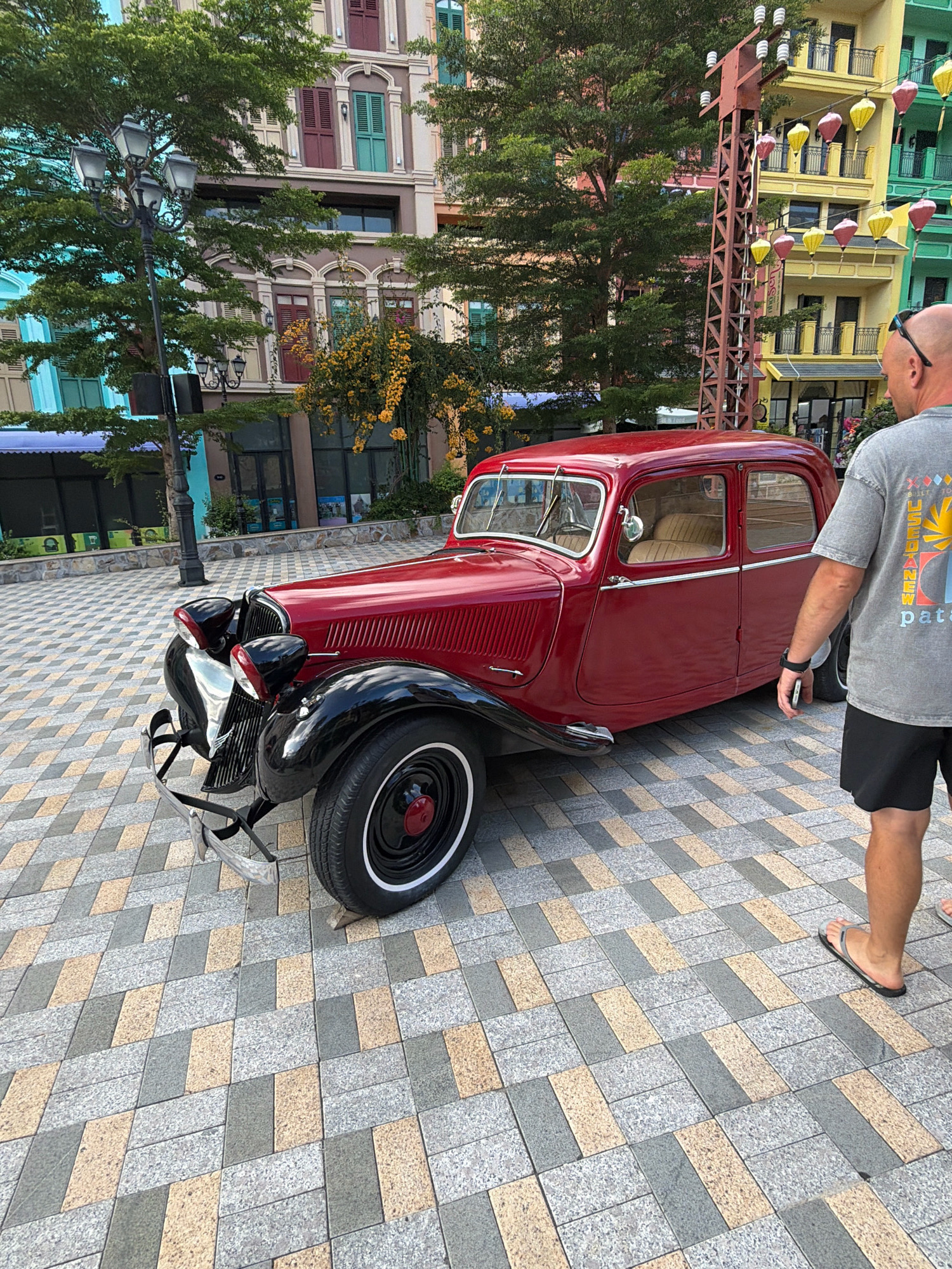 A man stands to the right of a vintage red Citroën Traction Avant car parked on a pedestrian walkway in Nha Trang, Vietnam. The man, wearing a grey t-shirt and black shorts, is viewed from the back, with his right arm bent at the elbow and his hand near his waist. He wears black flip-flops on his feet. The car, a four-door sedan, is positioned diagonally across the frame, with its front facing towards the left. The walkway is paved with a pattern of rectangular tiles in shades of grey, beige, and speckled grey. In the background, colorful buildings with balconies and shutters are visible, along with lush green trees and a decorative lamppost. Lanterns in shades of yellow, pink, and green hang from a metal structure. The sky is not visible.