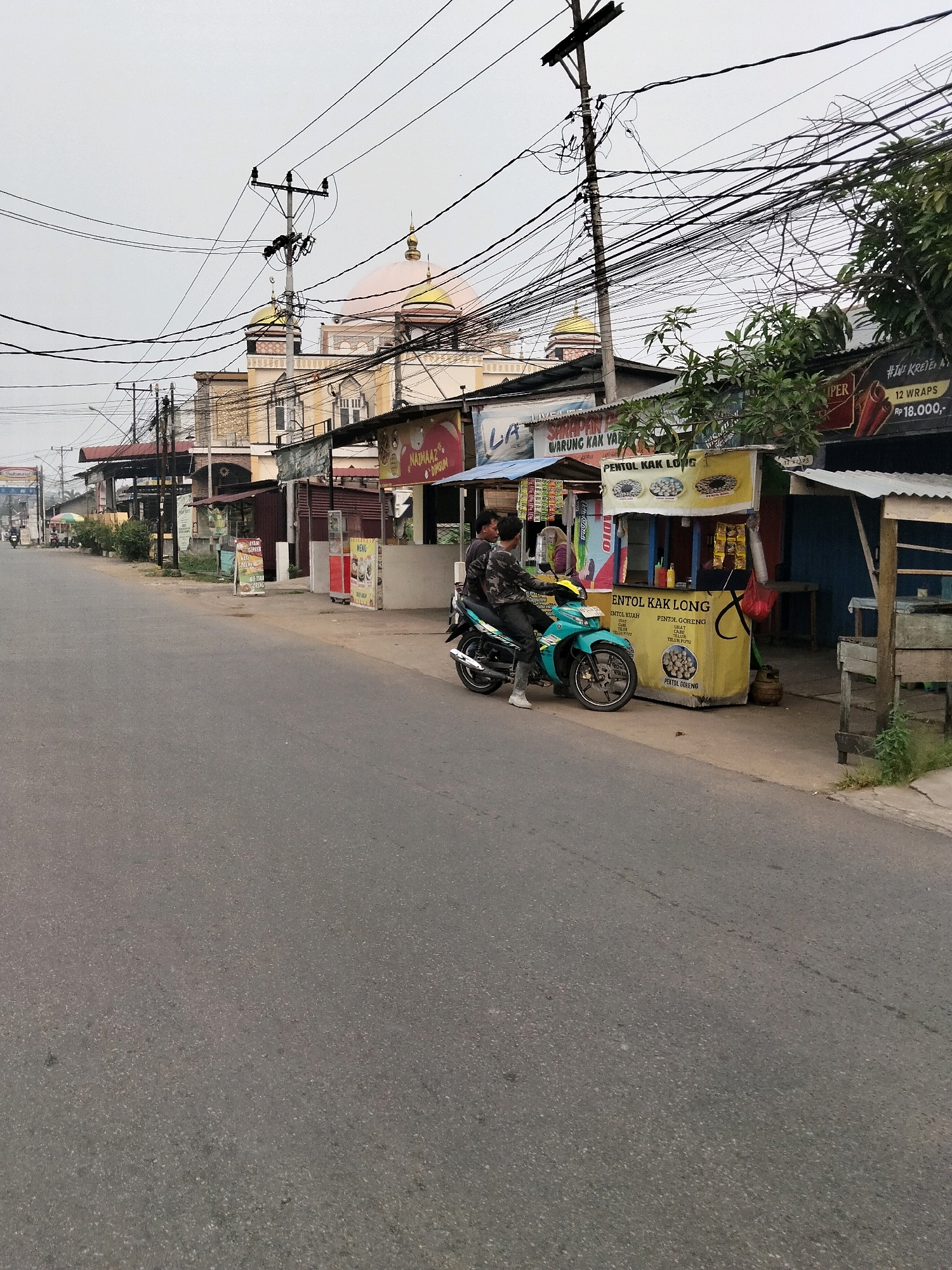 The image shows a street scene in the city of Arang Limbung, Indonesia, with a mosque in the background. A paved road occupies the foreground, with a sidewalk to the right. Along the sidewalk, several small businesses are visible, including food stalls and shops. One food stall, yellow in color, displays "PENTOL KAK LONG" signage and has a motorcycle parked in front of it. Two individuals are seated on the motorcycle, facing towards the stalls. Overhead, numerous power lines crisscross the sky, supported by utility poles. The mosque, a prominent structure, features a pale pink dome and arches. The sky is overcast, suggesting a cloudy day. The overall impression is of a bustling street with local commerce and religious architecture.