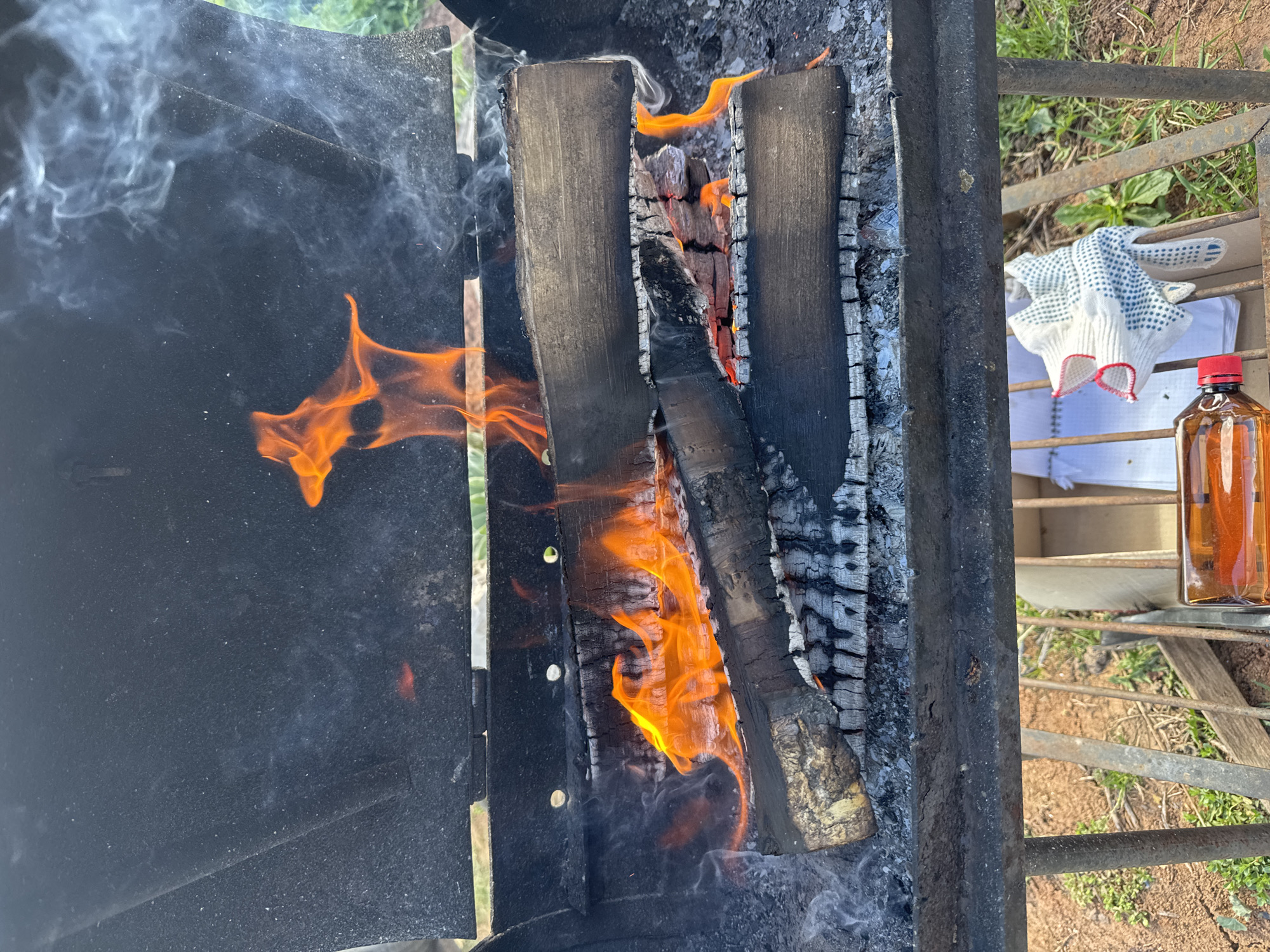 The image shows a grill with burning wood inside. The wood is charred and emitting flames. There is smoke rising from the fire. To the right of the grill, there are gloves, a sheet of paper, and a bottle of liquid, presumably for grilling. The background suggests an outdoor environment with grass and dirt. There is no information in the image that indicates this scene is in Liukshud'ya, Russia.