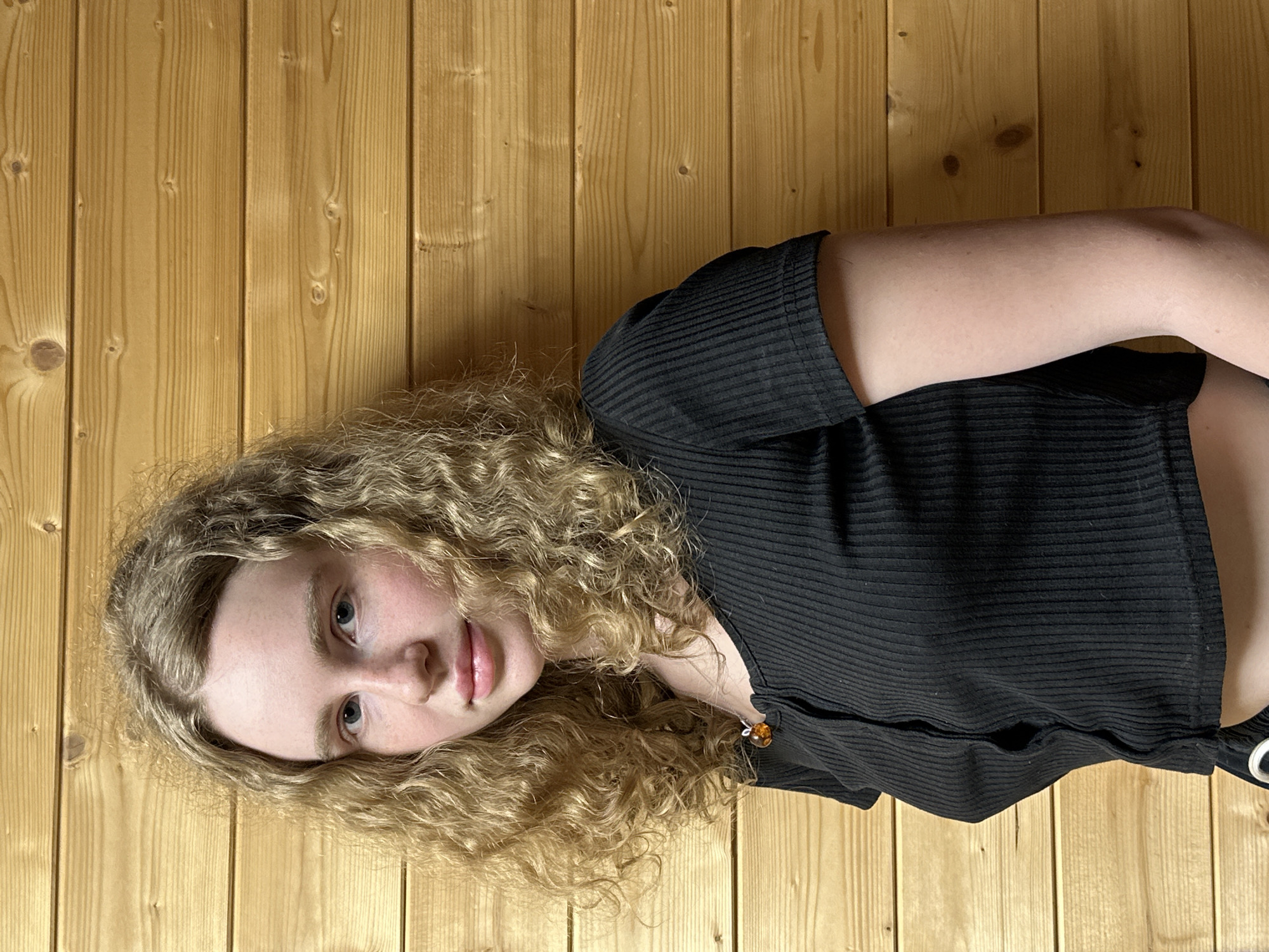 The image is a medium close-up portrait of a young woman, presented rotated 90 degrees clockwise. She has long, voluminous blonde curly hair that falls around her face and shoulders, appearing to rest against a wooden surface. Her light-colored eyes look directly at the camera with a calm, neutral expression, and her lips are slightly parted.
She is wearing a black, ribbed, short-sleeved top, which appears to be a crop top, with a small portion of her midriff visible. A small, shiny, amber-colored charm or decorative button is visible on the front of her top, near her collarbone. Her left arm is visible, bent at the elbow.
The background consists of vertical light-brown wooden planks, characterized by clear wood grain and several dark knots, suggesting an indoor setting with wood paneling. The lighting is soft and even, illuminating the scene without harsh shadows. There is no visible text in the image. The scene is located in Vasilyevo, Russia.