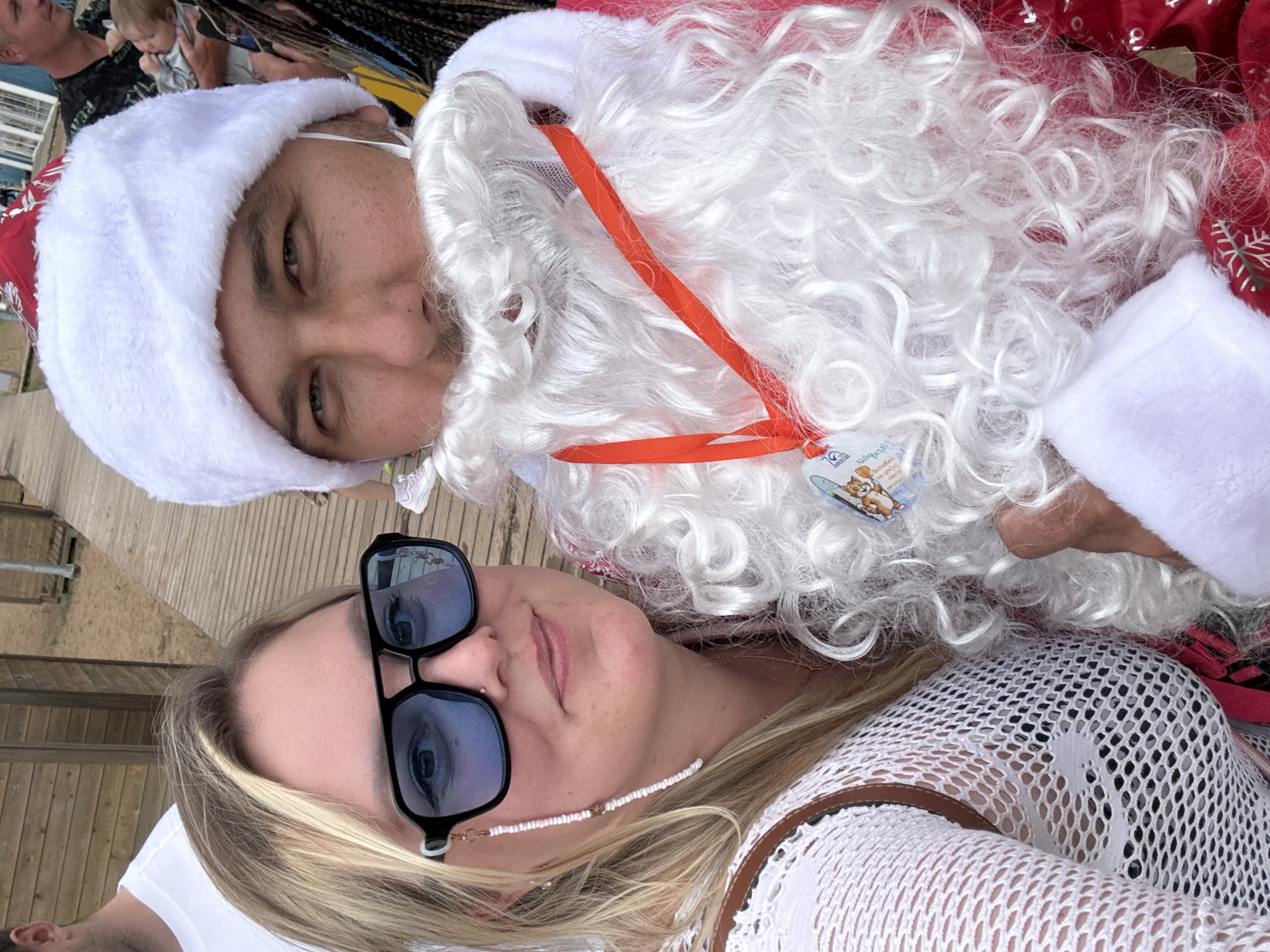 This media file features a close-up, overhead shot of two people, one dressed as Santa Claus. The person in the Santa costume, on the left, wears a white Santa hat and a white beard. They are looking down with their eyes partially closed. The second person, on the right, is wearing sunglasses and a white, netted top. They are looking up at the camera. Both individuals appear to be lying down.
In the background, out of focus, a baby can be seen being held by an adult. The scene appears to be outdoors, possibly on a wooden deck or boardwalk, suggested by the wooden planks visible. There are some metal railings to the left and a sandy area further in the background. The lighting suggests it is daytime. The overall mood seems relaxed and festive.
A small, colorful tag is visible on the Santa beard, with some text and an image of a cat. The text on the tag is partially obscured but appears to be in a foreign language, likely Russian given the location context of Chaikovsky, Russia. One visible word might be "KINDER" and another "сюрприз" which translates to "surprise." The person in the Santa costume has a red and white patterned fabric visible on their shoulder, consistent with a Santa suit. The person on the right has a thin, white beaded necklace with a small gold bead.