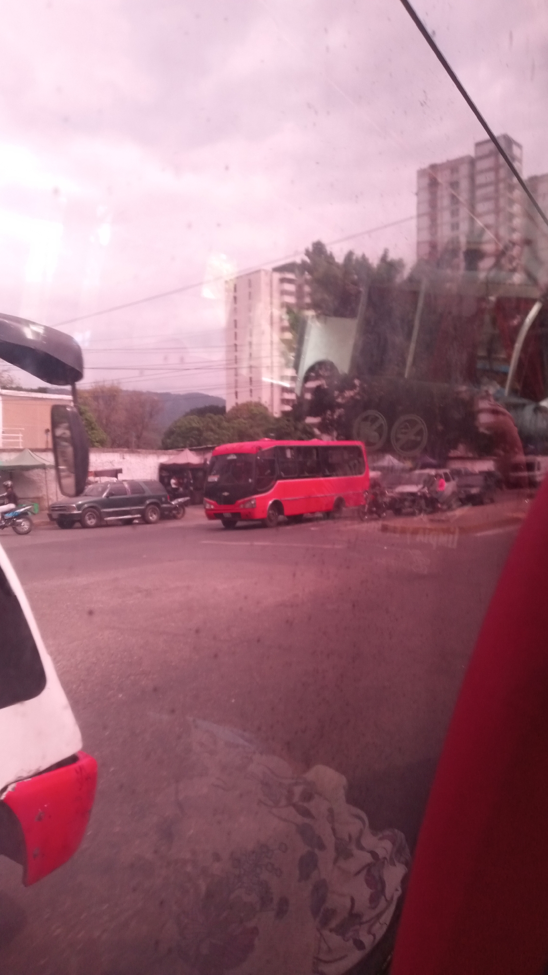 A red bus is on the street. It is a sunny day. This image seems to be taken from the inside of a bus. It is likely Caracas, Venezuela.