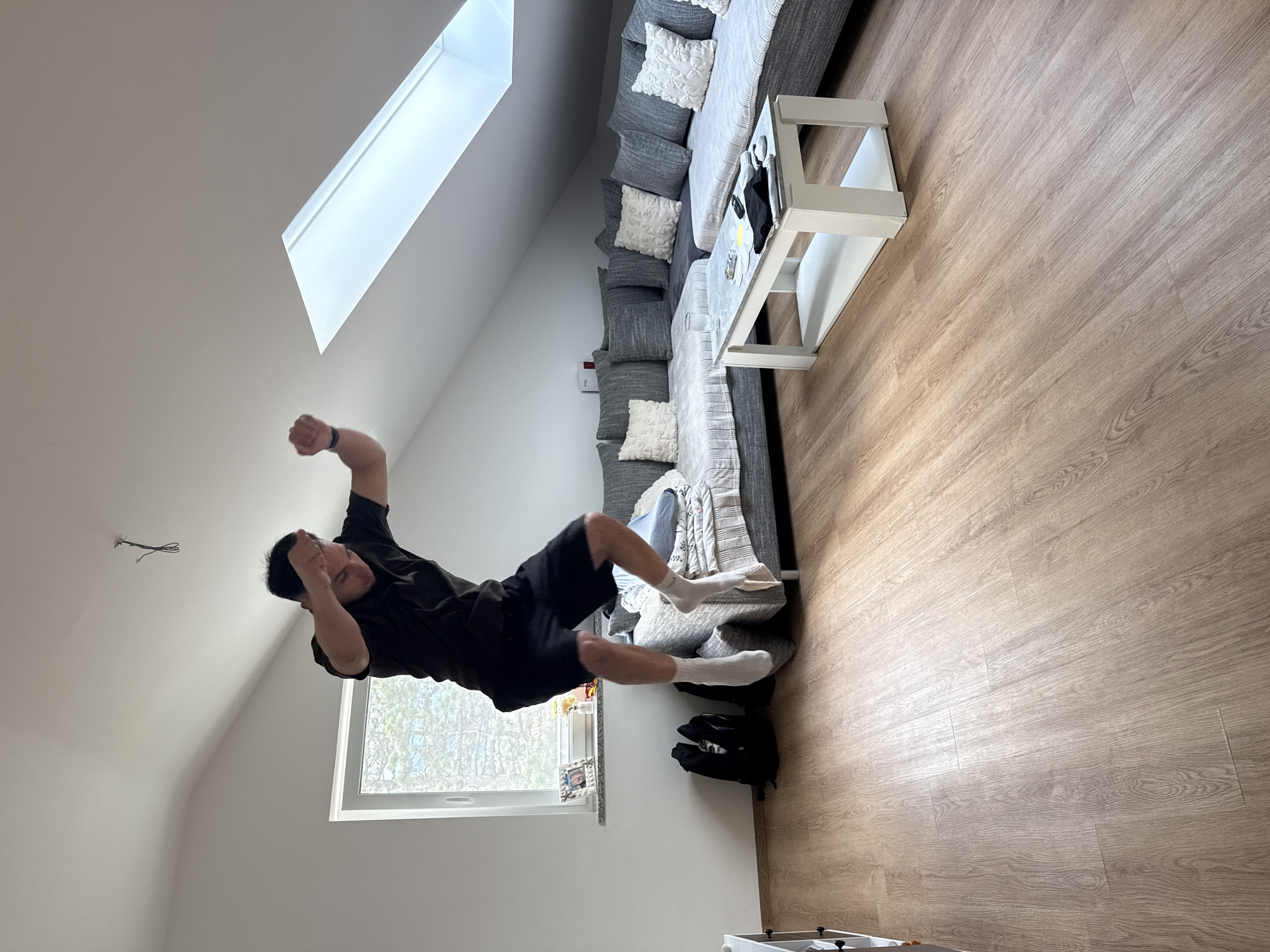 An adult male, wearing a dark t-shirt, dark shorts, and white socks, is captured mid-air in a jump within a residential attic room in the city of Garmisch-Partenkirchen, Germany. His arms are bent with fists clenched, and his facial expression suggests exertion or excitement. The room features white walls and a sloped ceiling, with light wood-patterned flooring. A large window on the left wall offers a view of outdoor trees, while a rectangular skylight is present on the right sloped ceiling section. Exposed electrical wires hang from a point on the main ceiling. An L-shaped sectional sofa, upholstered in grey and covered with light-colored throws and numerous grey and white pillows, occupies the back and right side of the room. A white rectangular coffee table with a lower shelf stands in the center foreground, holding a clear glass of water, a mobile phone, a folded black garment, a remote control, and a yellow circular object. Two black backpacks are visible on the floor near the window, adjacent to the sofa. A framed photograph, small plants, and other decorative items are arranged on the windowsill. A partial view of a white cabinet is visible on the far left.