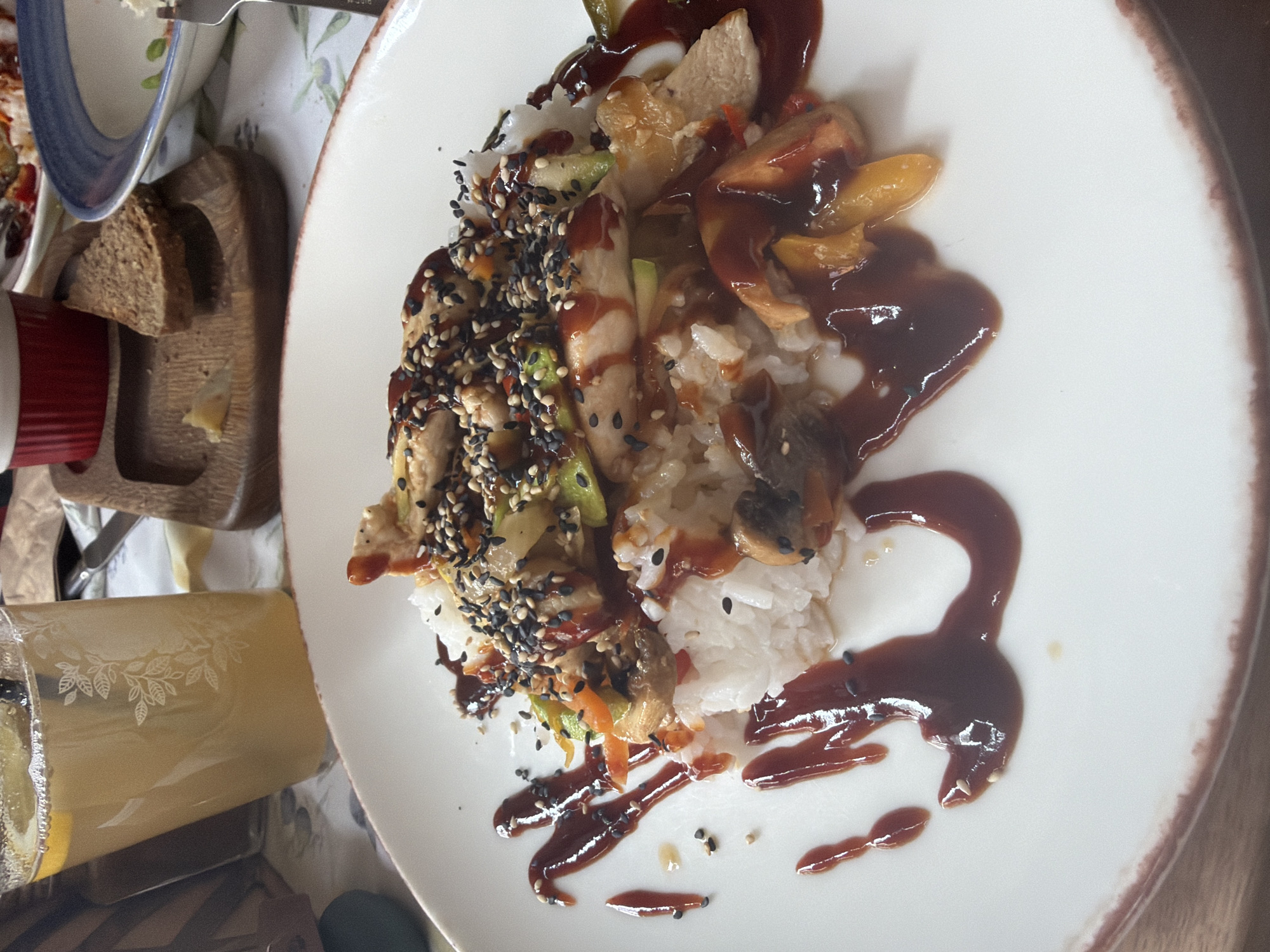 The image shows a plate of food, likely in a restaurant setting. The main dish consists of white rice topped with a mix of ingredients, including sliced chicken, sautéed mushrooms, and what appear to be stir-fried vegetables (possibly bell peppers, carrots, and zucchini). The dish is heavily garnished with black sesame seeds and drizzled with a dark, glossy sauce, likely a teriyaki-type sauce. The sauce is also used decoratively on the plate. To the left of the plate is a glass of lemonade with lemon slices visible inside. Further left, there are a small cutting board with slices of brown bread and a bowl, partially visible. The image is cropped at an angle. Location and city are not discernible from image.