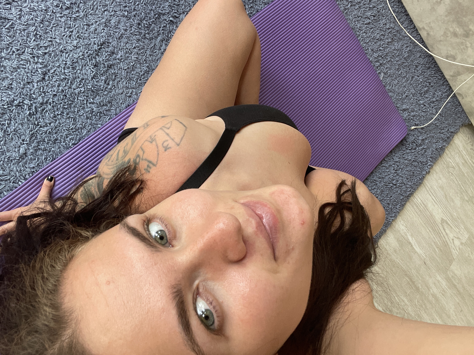 A high-angle close-up photograph depicts a woman lying on a purple yoga mat placed on top of a grey shag rug, situated on a light-colored wooden floor. She wears a black sports bra and her right shoulder has tattoos. The woman's eyes are open and looking towards the camera. A white cord rests on the wooden floor adjacent to the rug. The location is unknown and cannot be determined from the image.
