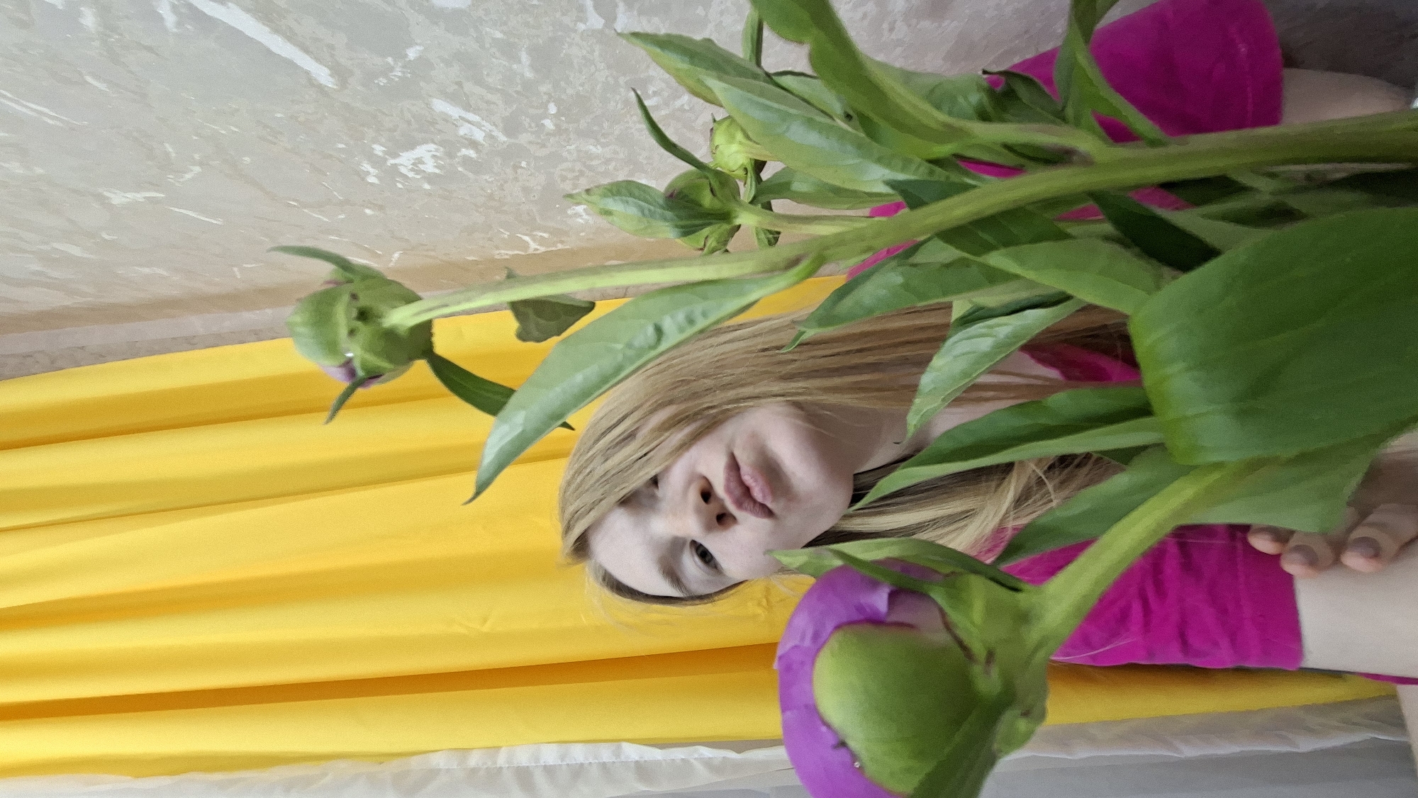 A young woman with blonde hair and a fuchsia top is looking directly at the camera, her face partially obscured by green leaves and flower buds. She has a neutral expression, with her lips slightly parted. The image is taken at a high angle, looking down on her, and is rotated on its side, making the yellow fabric behind her appear as a vertical background. One large, green flower bud with a hint of purple at its base is prominent in the foreground, bottom right. Another smaller, entirely green bud is visible in the top left. The background consists of a crumpled yellow fabric, possibly a curtain or tablecloth, behind the woman, and a textured light-colored surface, perhaps a wall or ceiling, above it. The overall impression is an intimate, close-up shot focusing on the woman and the vibrant plant. No visible text is present.