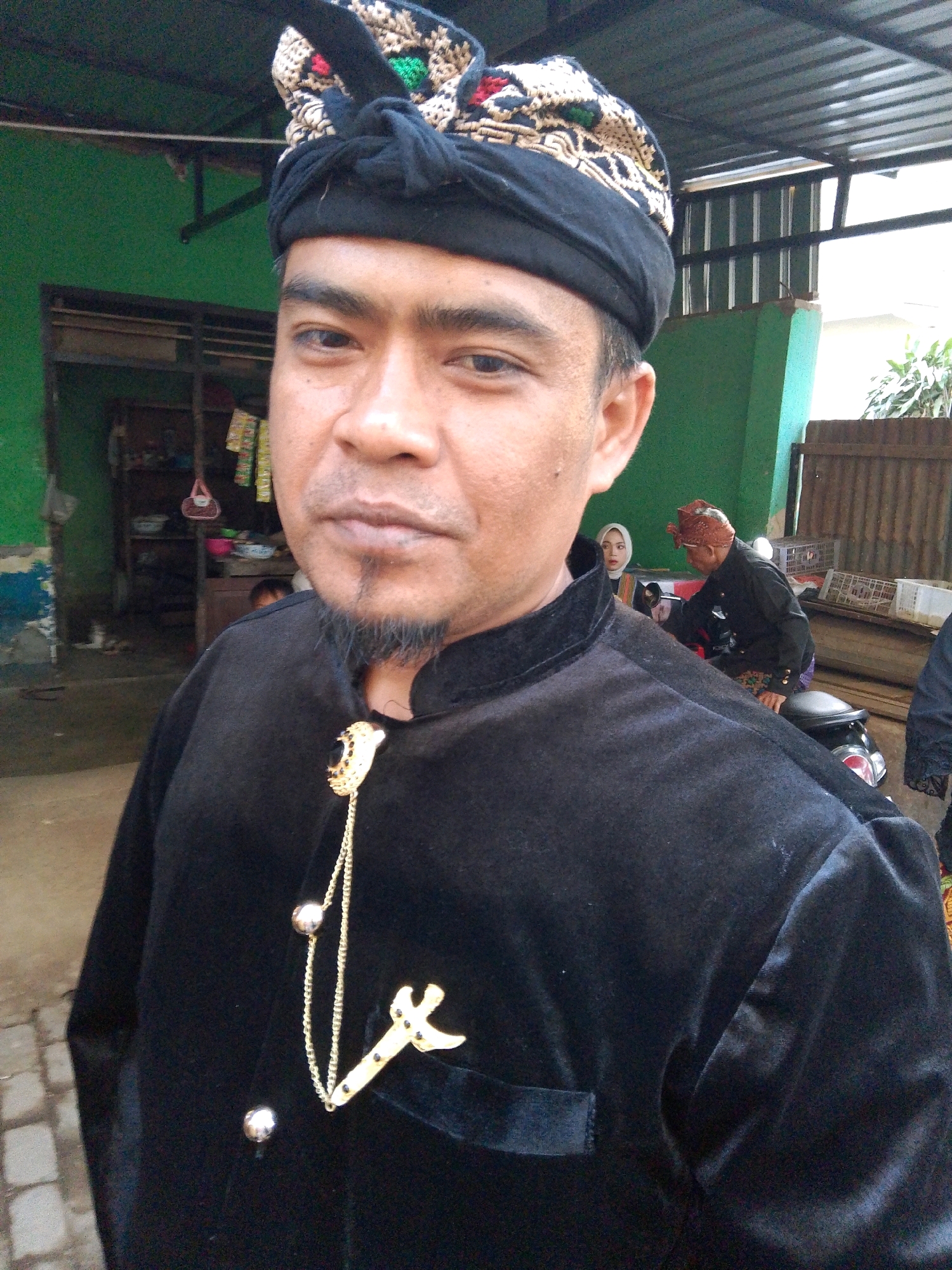 A man wearing a black velvet jacket and a patterned head covering is pictured in an outdoor setting that appears to be in Kuripan Utara, Indonesia. The man has a goatee and is looking towards the camera with a neutral expression. He is adorned with a gold-colored chain and emblem on his jacket. In the background, there is a green wall, a wooden fence, and other individuals partially visible. One person is sitting on a motorcycle, and another person wearing a hijab is standing. The scene suggests a cultural event or gathering.