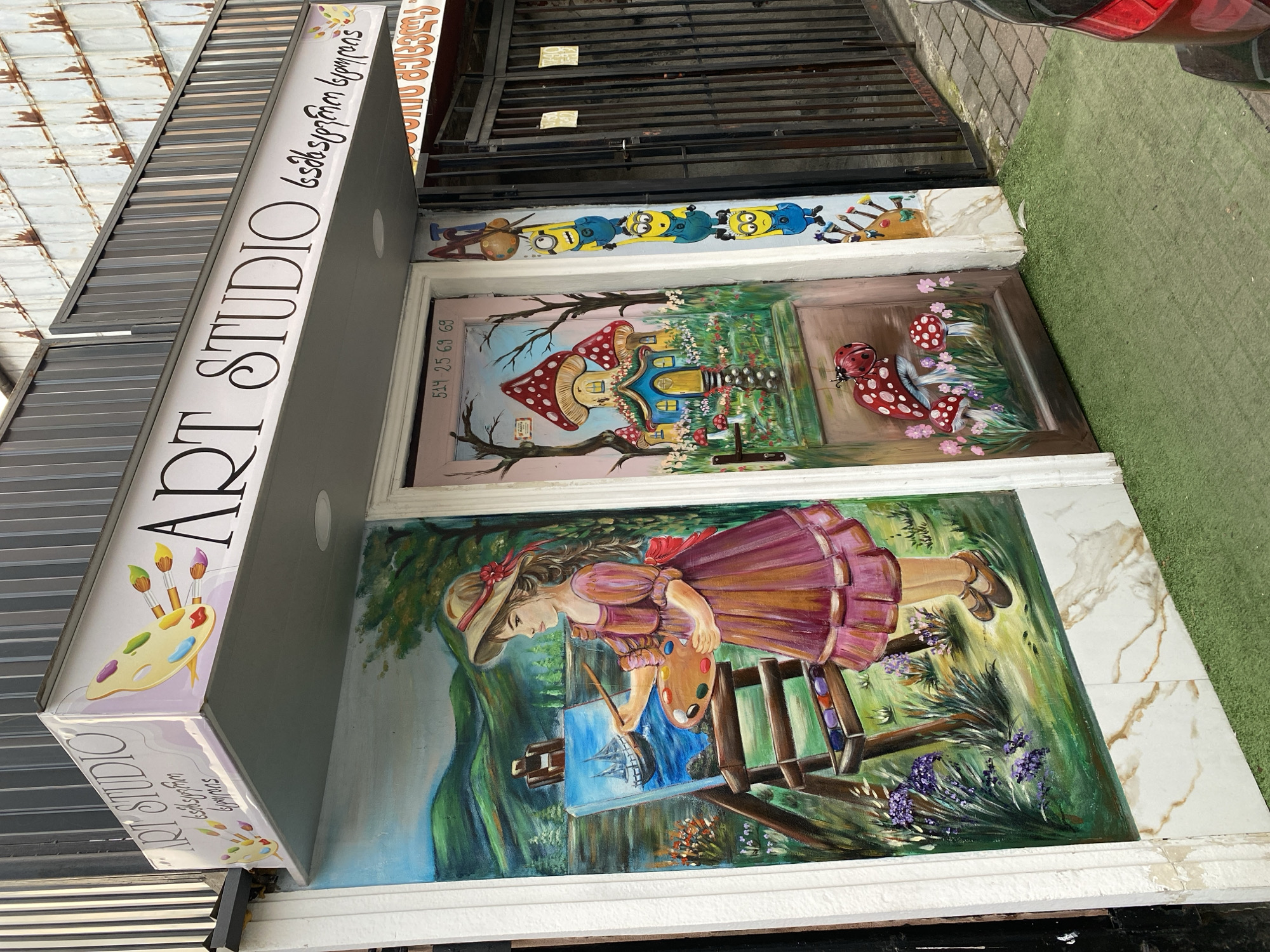 This is a photograph of the exterior of an "ART STUDIO" located on a street in Batumi, Georgia. The studio features a colorful mural on its facade, depicting a young girl in a pink dress painting a sailboat on an easel. The mural extends to a door painted with a whimsical scene of mushroom houses and ladybugs. The studio's signage is visible above the entrance, with "ART STUDIO" written in English and Georgian. To the right of the door, a column is decorated with cartoon characters resembling Minions. The street in front of the studio is paved with cobblestones, and artificial turf is laid out as a pathway. A portion of a car is visible in the lower right corner of the image. The building appears to be situated in an urban environment, with a corrugated metal wall and a gate visible in the background.