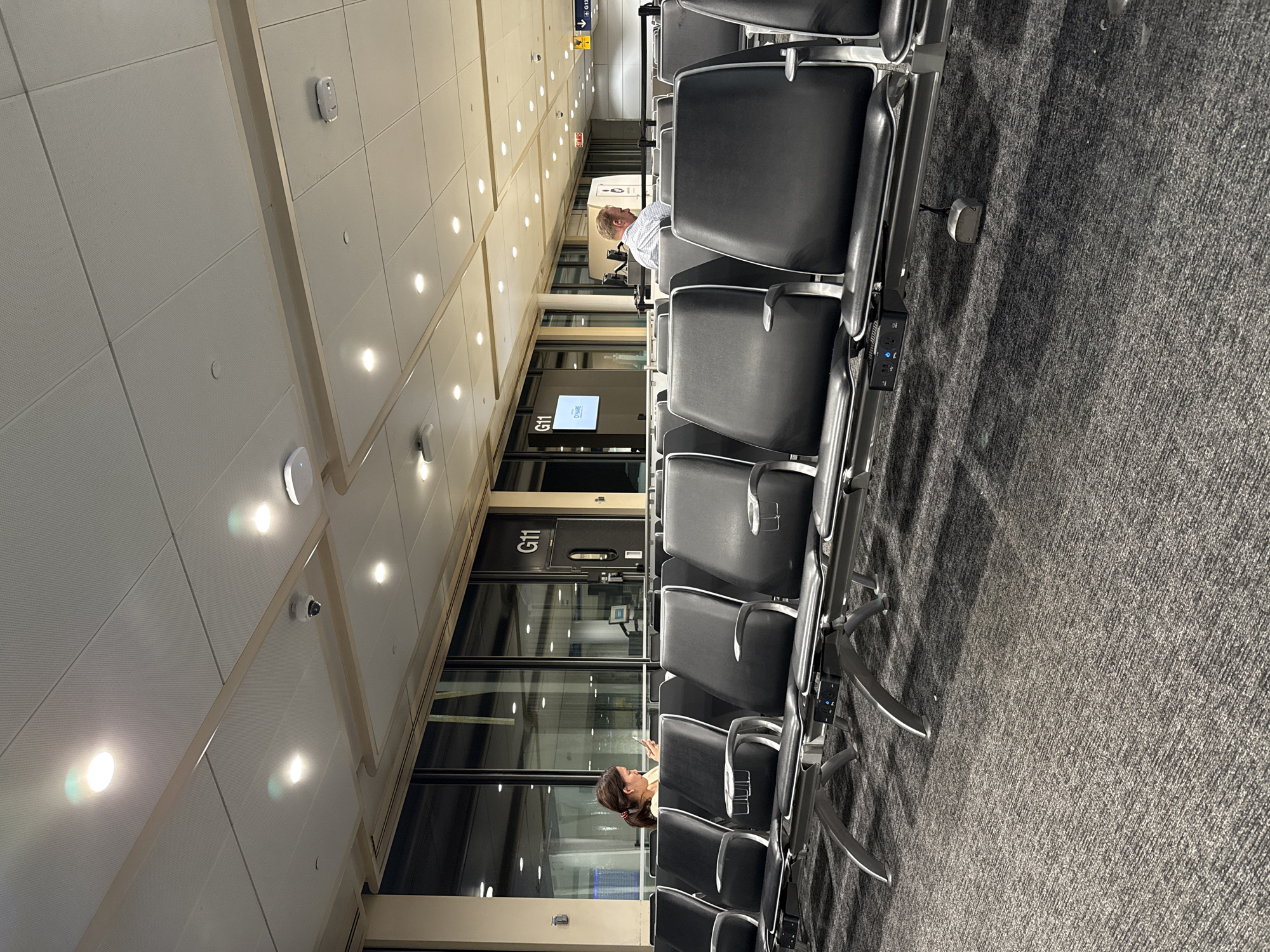 The image depicts an airport terminal waiting area. Rows of black chairs with metal frames line the walkway, which is covered in gray carpeting. Passengers occupy some of the seats, one of whom is looking down at a mobile device, and another person further down. The environment suggests a waiting area for Gate G11, as seen on a digital display on the nearby glass. Overhead, the ceiling is composed of rectangular panels with recessed lighting fixtures and surveillance cameras. The environment, while not explicitly confirmed, is likely inside of the Chicago O'Hare International Airport (ORD) due to the presence of Gate G11, which is located in this airport.