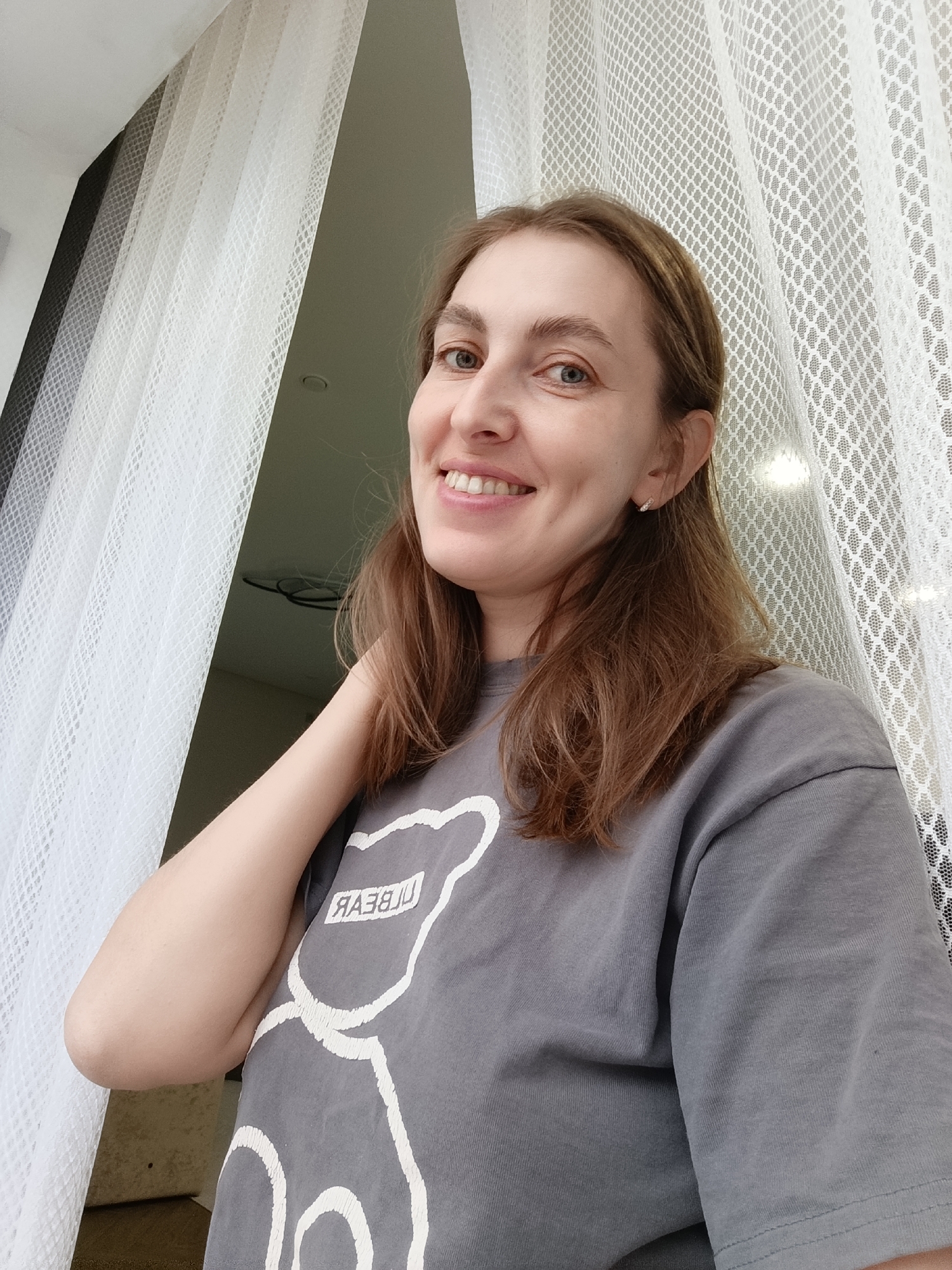 A woman with light brown hair, fair skin, and blue eyes smiles at the camera. She is wearing a gray t-shirt with a white outline of a bear graphic and the word "BEAR" in white reverse text. She has a small silver earring in her left ear. Her left arm is bent and her hand is resting on her neck. She is positioned in front of a window with sheer white curtains. The background appears to be an indoor setting with a ceiling visible above the curtains. The lighting suggests it is daytime. The image was taken in Chaikovsky, Russia.