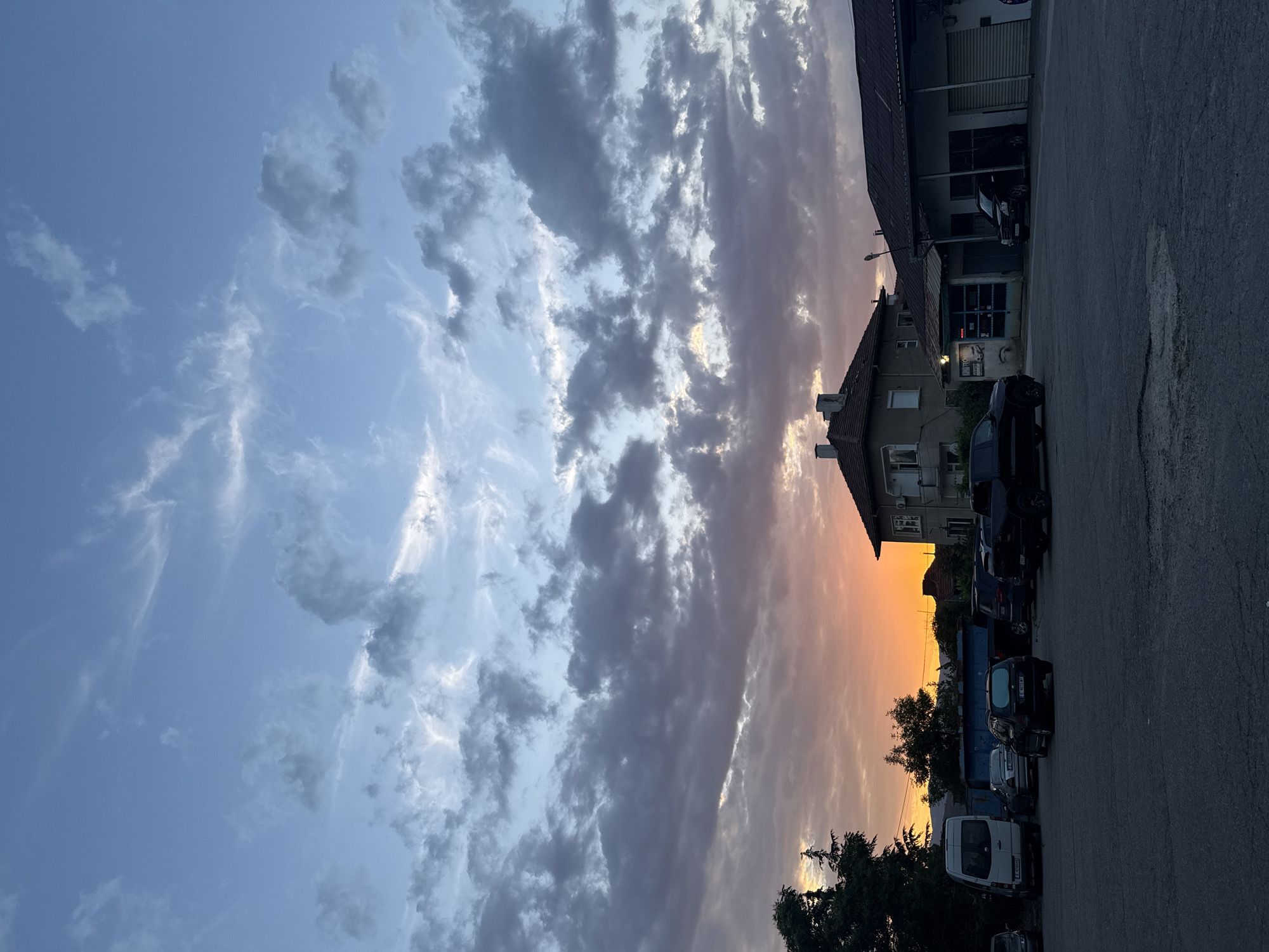 The image captures an outdoor scene in Blagoevgrad, Bulgaria, at sunset. The sky displays a clear gradient from light blue at the zenith, transitioning into dense, dark grey clouds streaked with white and tinged with pink and orange hues towards the horizon. The setting sun casts a vivid orange and yellow glow along the lower sky, visible through gaps in the cloud cover. The foreground consists of a dark asphalt surface, likely a parking lot, illuminated dimly.
On the left side, a dark green coniferous tree and another deciduous tree are prominent. A white panel van (identifiable by a Bulgarian license plate starting with 'E' for Blagoevgrad) is parked facing away from the viewer. Behind it, additional vehicles are partially obscured.
In the midground, a two-story residential building with a brown tiled roof and multiple chimneys stands on the left. Its facade is a light, neutral color. To its right, a single-story building, possibly a workshop or garage, features a dark roof and an open bay with a dark vehicle parked inside. Several other dark-colored vehicles, including a station wagon (with a Bulgarian 'E' plate) and a large pickup truck, are parked in the lot. A blue industrial skip or container is visible between some of the parked vehicles. No people are discernible in the scene. The overall lighting suggests dusk, with the ground and structures in shadow against the brightly colored sky.