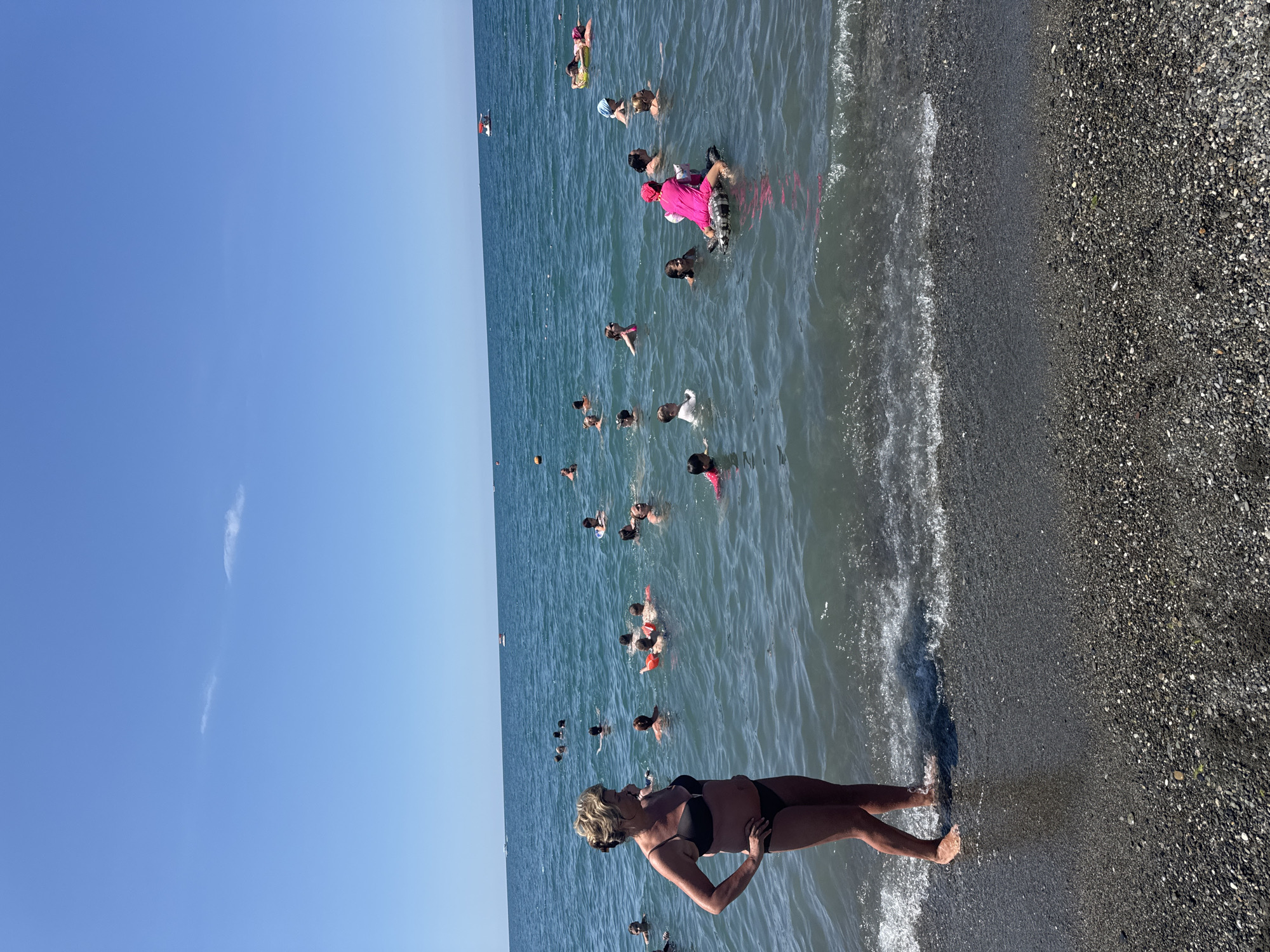 The image depicts a crowded beach scene with numerous people swimming in the Black Sea off the coast of Sochi, Russia, on a clear, sunny day. In the foreground, a woman stands at the water's edge, facing away from the camera. The water is calm with gentle waves lapping the pebbly shore. Further out, a diverse group of individuals, including adults and children, are engaged in various water activities. Some are swimming or wading, while others are floating on inflatable toys. In the distance, two boats are visible on the horizon, and a few more people are scattered across the water. The sky is a vibrant blue with a few wispy clouds. The overall atmosphere suggests a lively and enjoyable day at the beach.