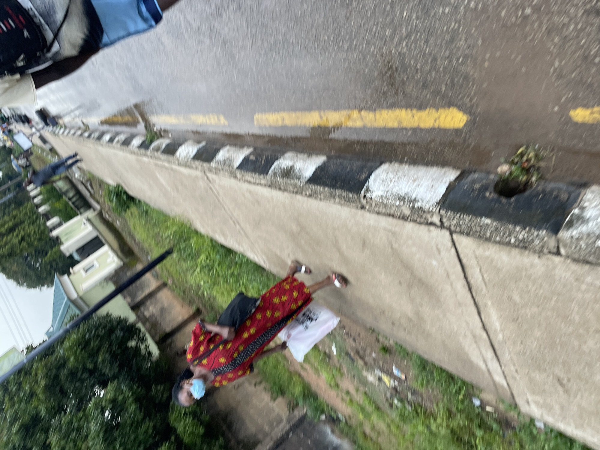 A woman wearing a face mask and a red and yellow patterned dress walks on a sidewalk beside a road in Jos, Nigeria. The sidewalk is cracked and overgrown with grass in places. The road is wet, with yellow lines painted on it. There are some buildings visible in the background, as well as trees and greenery. The woman is carrying a black bag over her shoulder and holding a white plastic bag in her hand. Another person, wearing blue shorts and a patterned shirt, is walking away from the camera on the road. The scene appears to be a typical urban environment in Jos, with evidence of recent rain.