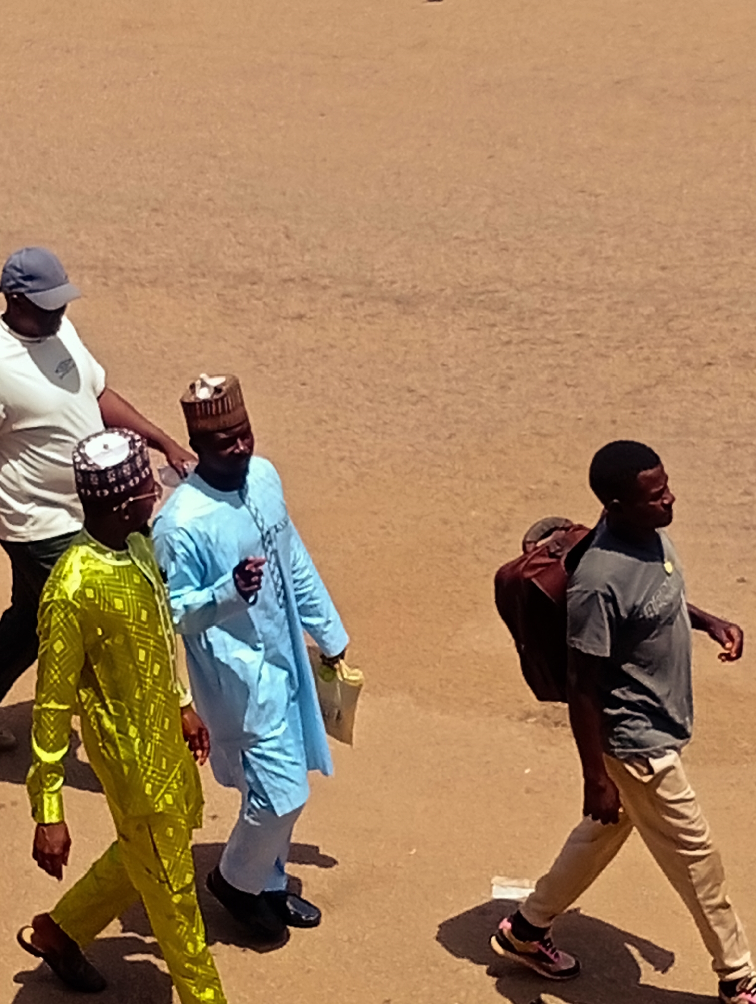 In Bukuru, Nigeria, four individuals are captured in a high-angle shot walking across a sun-baked, dirt surface. On the left, a person wearing a blue baseball cap and a white t-shirt walks away from the camera. To their right, two men dressed in traditional attire are walking side-by-side. The man in the foreground is wearing a bright yellow, intricately patterned robe and pants, with a matching cap. The man next to him is in a light blue robe and pants, also wearing a patterned cap. He is gesturing with his right hand. Behind them, a fourth individual, a man in a grey t-shirt and tan pants, walks with a dark brown bag slung over his right shoulder. The ground appears to be a dry, dusty, unpaved area.