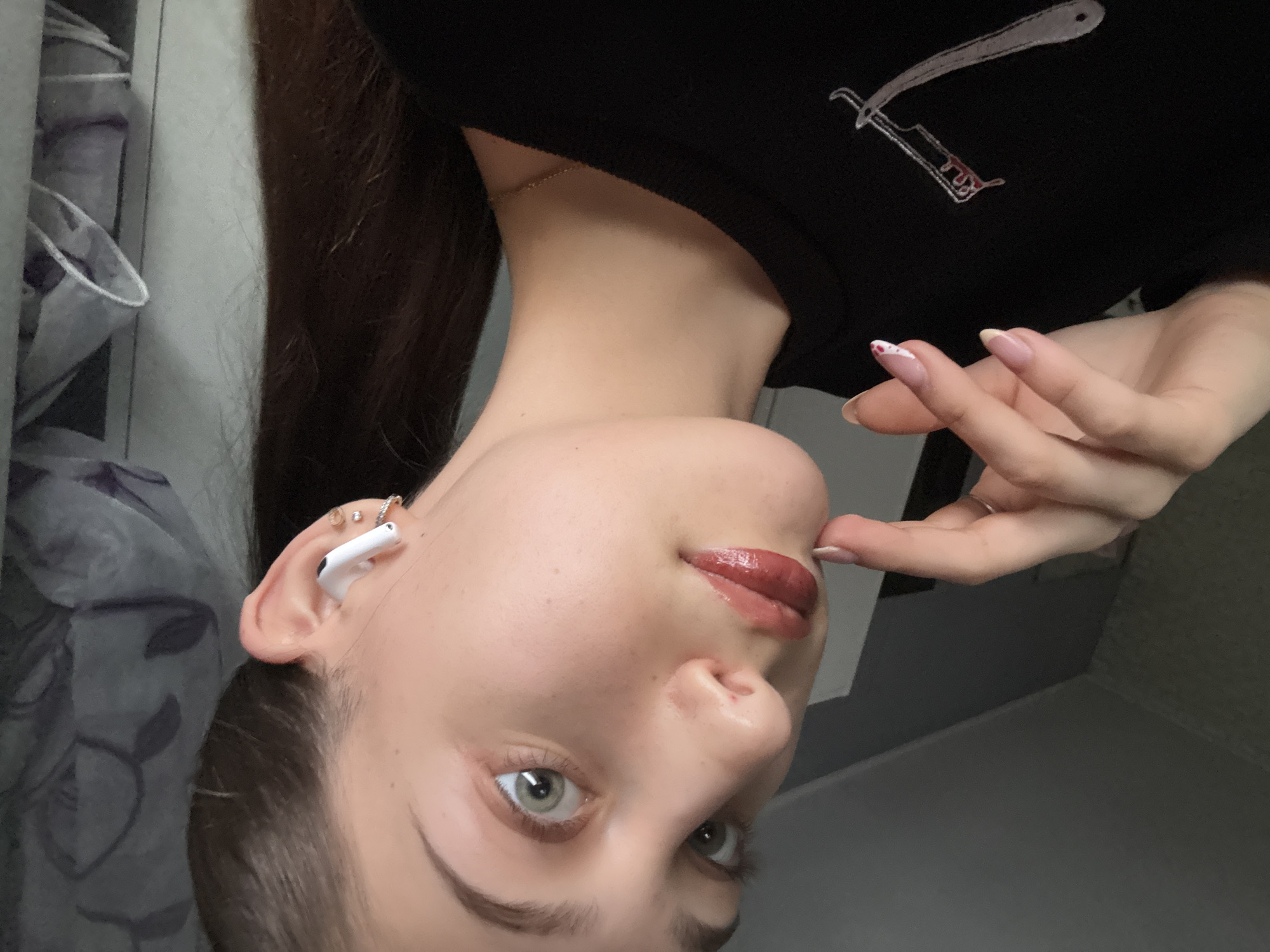 A selfie image shows a female individual with fair skin, light green eyes, and light brown eyebrows. She has dark brown hair pulled back and is wearing a black long-sleeved shirt with a graphic design on the upper right. Her left ear has multiple piercings and a white wireless earbud. Her nails are manicured in a French style with a heart design. Her lips have a glossy finish. The background includes a gray patterned curtain on the left and a gray wall on the right. The location is not specified. The image is rotated.