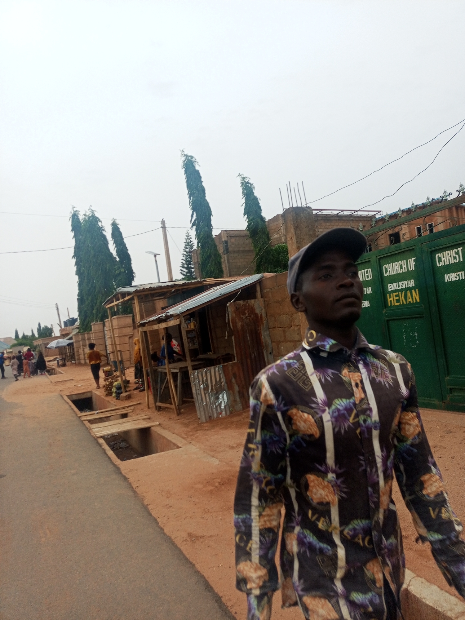 A photograph taken at street level depicts a man in the foreground. He is wearing a patterned shirt and a baseball cap. Behind him, a green gate reads "UNITED CHURCH OF CHRIST, EKKLISIYAR HEKAN, NIGERIA." Further into the background, there is a small, open-air shop structure. People are walking in the distance. The road is reddish-brown. There are open drainage ditches along the road. Tall, thin trees stand along the side of the street. The sky is overcast. The location is Tempure, Nigeria.
