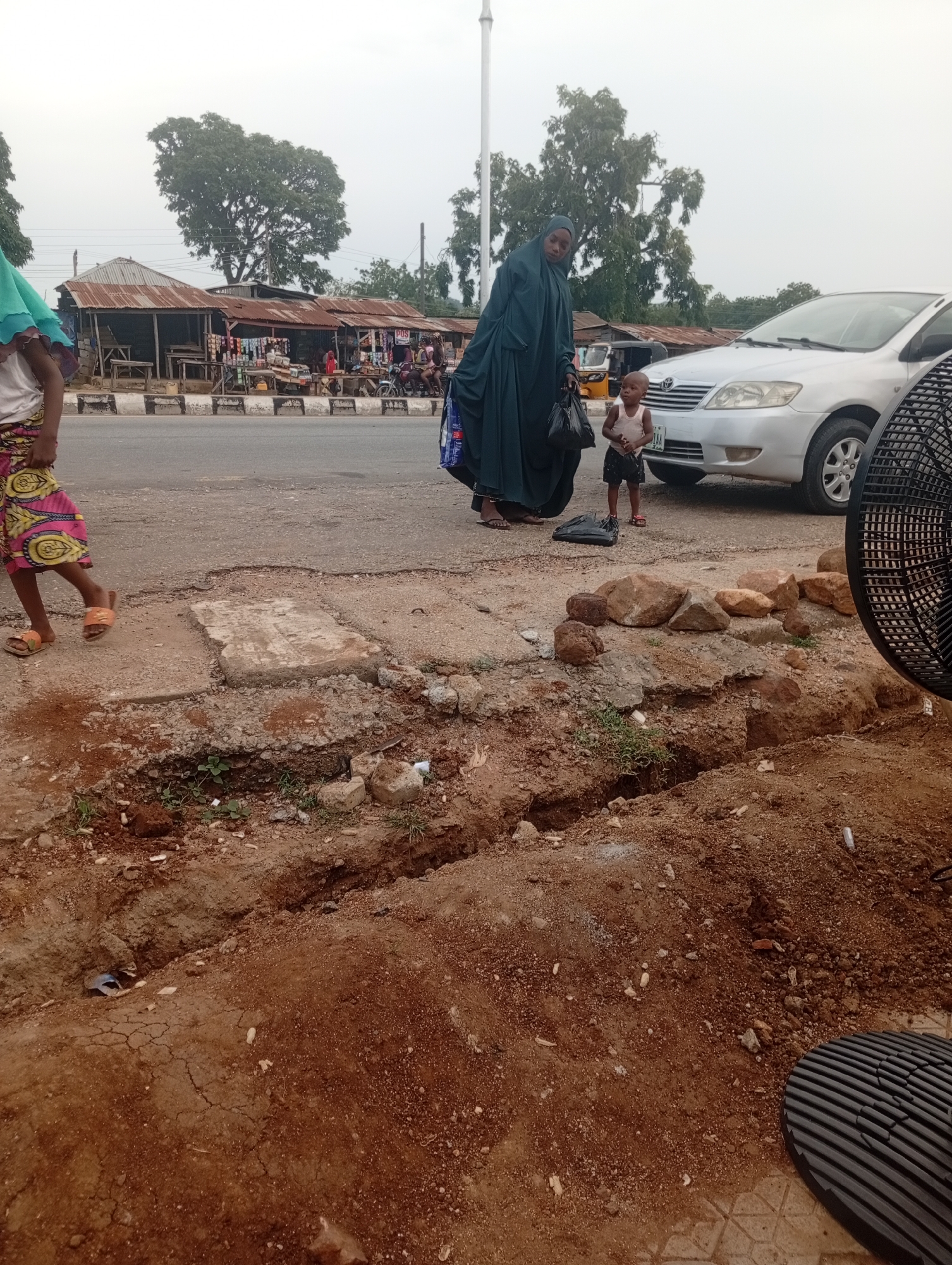 The image depicts a street scene in Zango-ndaji, Nigeria, with a focus on the foreground. In the foreground, a trench has been dug in the reddish-brown earth, exposing layers of soil and some scattered rocks and debris. A partially visible black, ribbed object, possibly part of a fan or grating, occupies the lower right corner of the frame.
Behind the trench, a paved road runs horizontally across the image, marked with white and black lines. On the left side of the road, a young girl in a colorful patterned skirt and orange sandals is walking away from the camera. To her right, a woman in a dark green hijab and long dress is standing, holding a black plastic bag. Beside the woman and slightly behind her is a young child wearing a light-colored tank top and dark shorts.
Across the road, a row of small shops with corrugated iron roofs are visible. The shops appear to be selling various goods, with shelves and displays lined up. A silver sedan is parked on the road, to the right of the shops and the people. The background is filled with trees and a hazy, overcast sky, suggesting it may be early morning or late afternoon. The overall scene captures a typical street environment with elements of commerce, daily life, and infrastructure work.