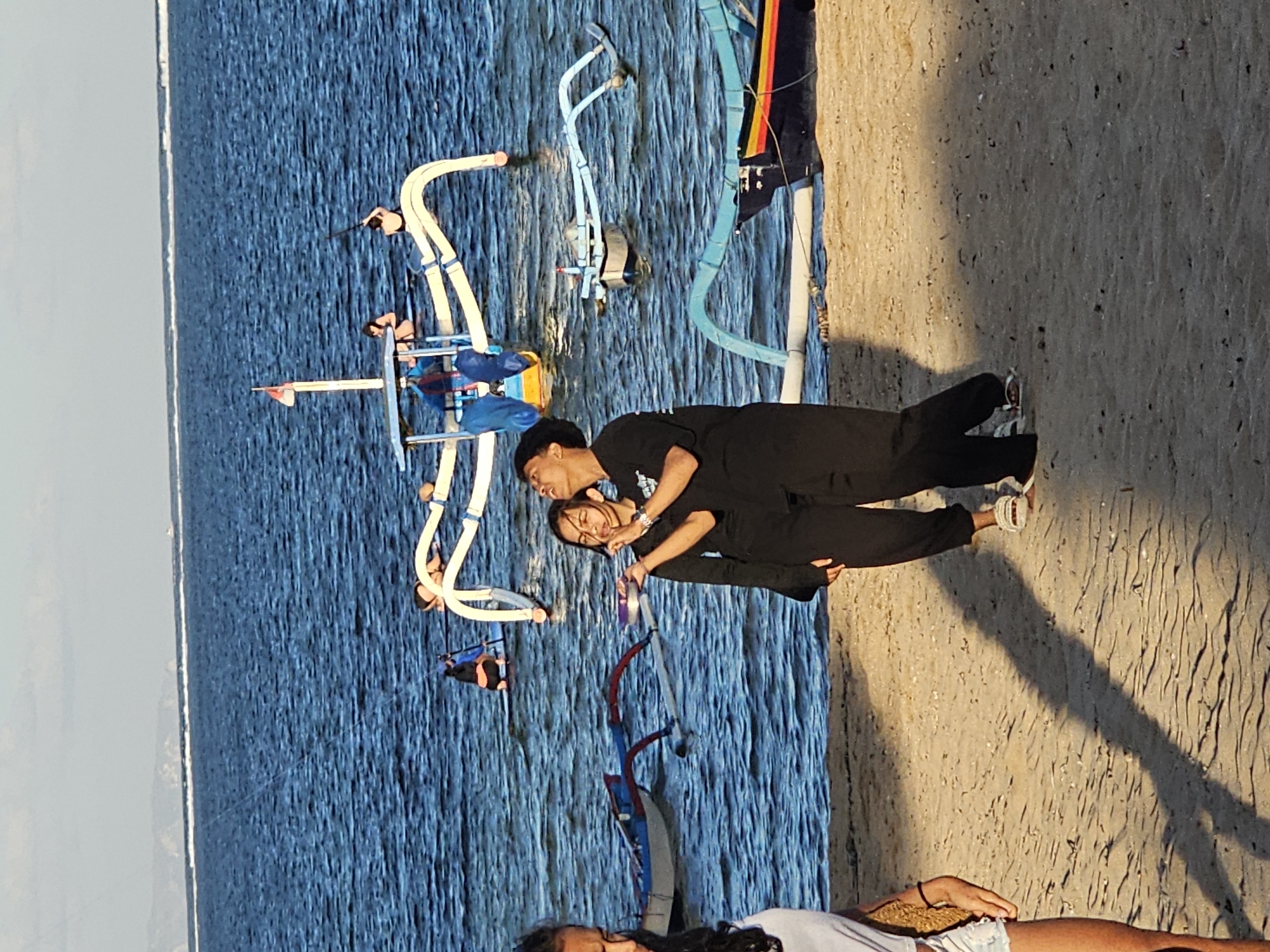 The image captures a serene beach scene in Denpasar, Indonesia, likely during the late afternoon given the soft, warm lighting and the long shadows cast on the sand. The foreground features a sandy beach with two individuals, a young man and woman, standing close together and looking up towards the sky. They are dressed in matching black outfits, suggesting they might be together. The woman is holding a small, colorful bowl.
In the background, the blue ocean stretches out, calm and inviting. Several traditional outrigger boats are visible, some moored near the shore, others further out in the water. One boat, centrally located, has a white frame with blue accents and a red and white flag. There are other people visible on or near these boats, some appearing to be engaged in activities on the water. The water exhibits gentle ripples, and in the far distance, a hazy coastline with hills can be seen under a clear sky. The overall atmosphere is peaceful and indicative of a relaxed day at the beach.