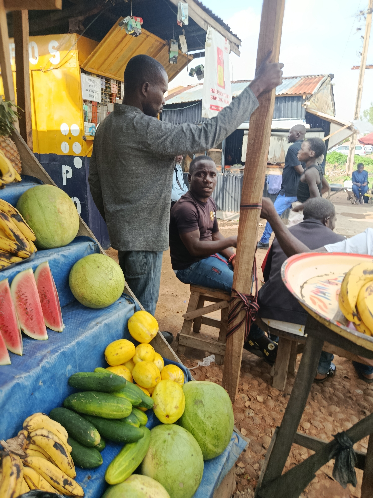 A street market scene is depicted in Jos, Nigeria, with an emphasis on produce. In the foreground, a variety of fruits and vegetables are displayed on blue tarpaulin-covered shelves. These include several large watermelons, bunches of bananas, slices of watermelon, numerous cucumbers, and piles of yellow melons, possibly pawpaw or a variety of melon. A man wearing a grey long-sleeved shirt and blue jeans stands behind the produce, his back partially to the camera, holding a wooden pole. To his right, another man in a brown t-shirt and blue jeans sits on a wooden stool, looking towards the camera. In the background, several other individuals are visible. Some are standing, while others are seated, suggesting a bustling market environment. The market appears to be set up outdoors, with a corrugated iron roof structure visible, characteristic of informal market stalls. The ground is dirt and covered with small stones. A yellow kiosk with white lettering is present in the mid-ground, with signage that is partially legible, including "OPEN YOUR ACCOUNTS HERE." Other market stalls and structures made of wood and corrugated iron are visible further back, along with some vehicles in the distance, indicating the urban setting of Jos.