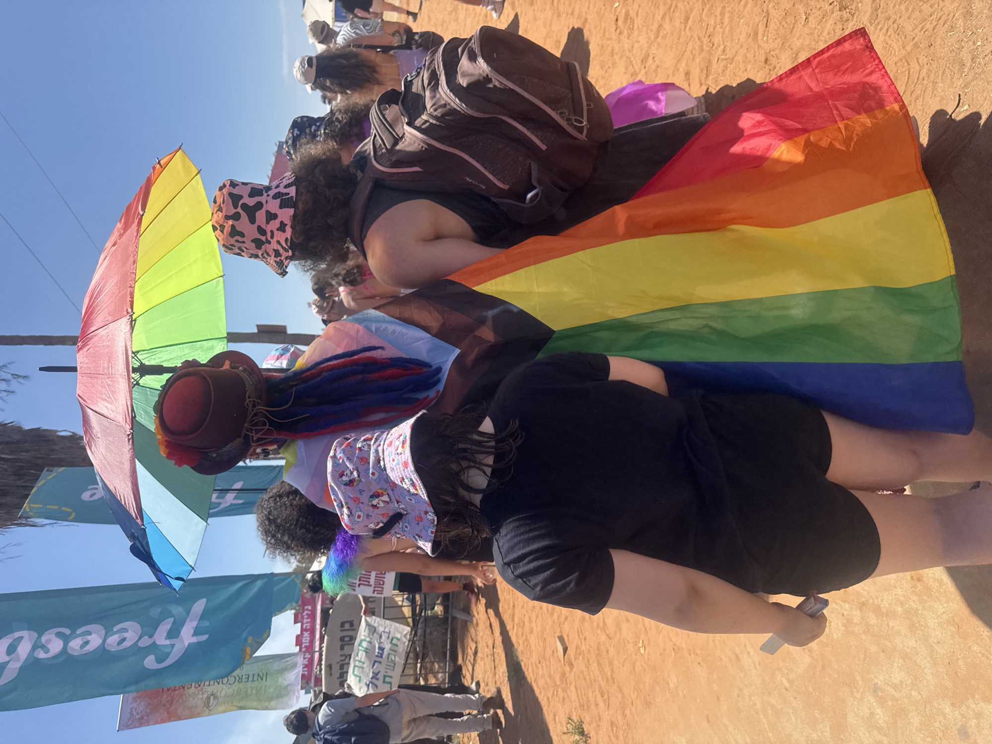 The image depicts an outdoor event, likely a parade or gathering, with a focus on LGBTQ+ pride. Several individuals are visible, most of whom are facing away from the camera. A prominent rainbow flag is draped over the back of a person in the foreground, who is wearing a black t-shirt and black shorts. Another person, also facing away, is wearing a bucket hat with a cow print pattern and has a backpack. Above them, a colorful umbrella provides shade. In the background, various flags, including a blue one with white text, are visible. There are other people in the background, some of whom are walking. The ground is covered in orange sand or dirt. Based on the style of flags and the overall atmosphere, this scene is consistent with events that take place during Tel Aviv Pride.