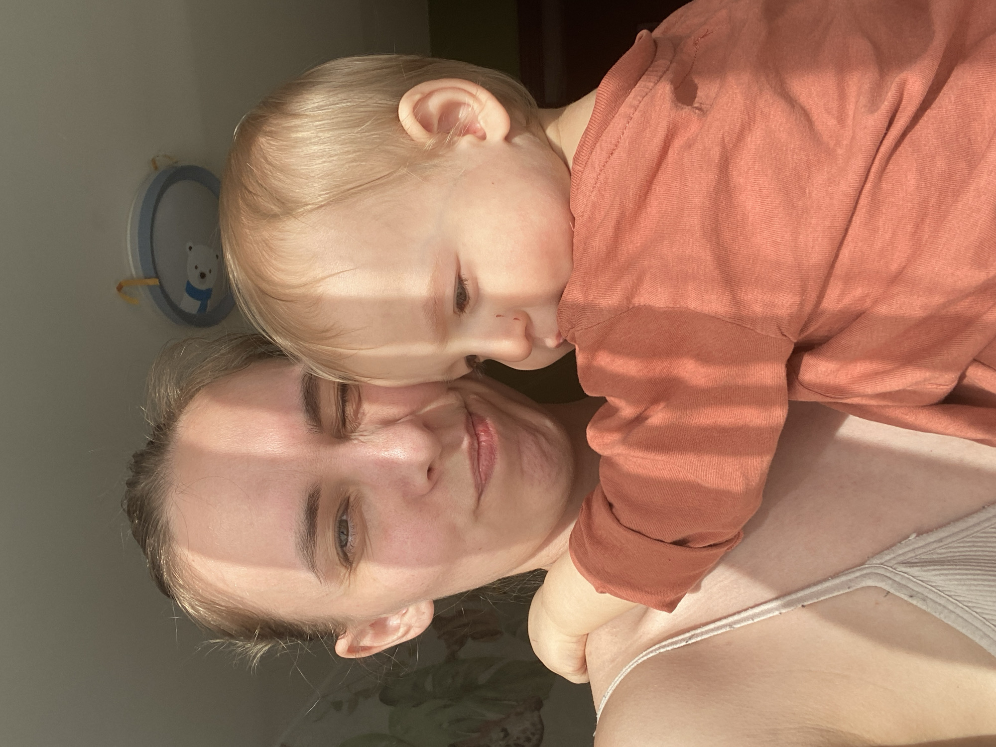A fair-skinned woman and a young child are in a close embrace. The woman is reclined, with her head resting on what appears to be a surface. She has blonde hair pulled back and is wearing a light-colored ribbed tank top. The child, with fair skin and blonde hair, is lying on top of her, face against the woman’s cheek. The child is wearing a salmon-colored long-sleeved shirt. Light streams in, casting shadows across the woman’s face and the child’s arm.
A round wall decoration, with a white bear design on a blue background, is partially visible in the upper left corner of the frame. The background includes sections of wall in varying shades of gray and glimpses of green and dark brown in the upper center.
The image was likely taken indoors during daylight hours. It's not possible to confirm the location in the city of Chaikovsky, Russia.