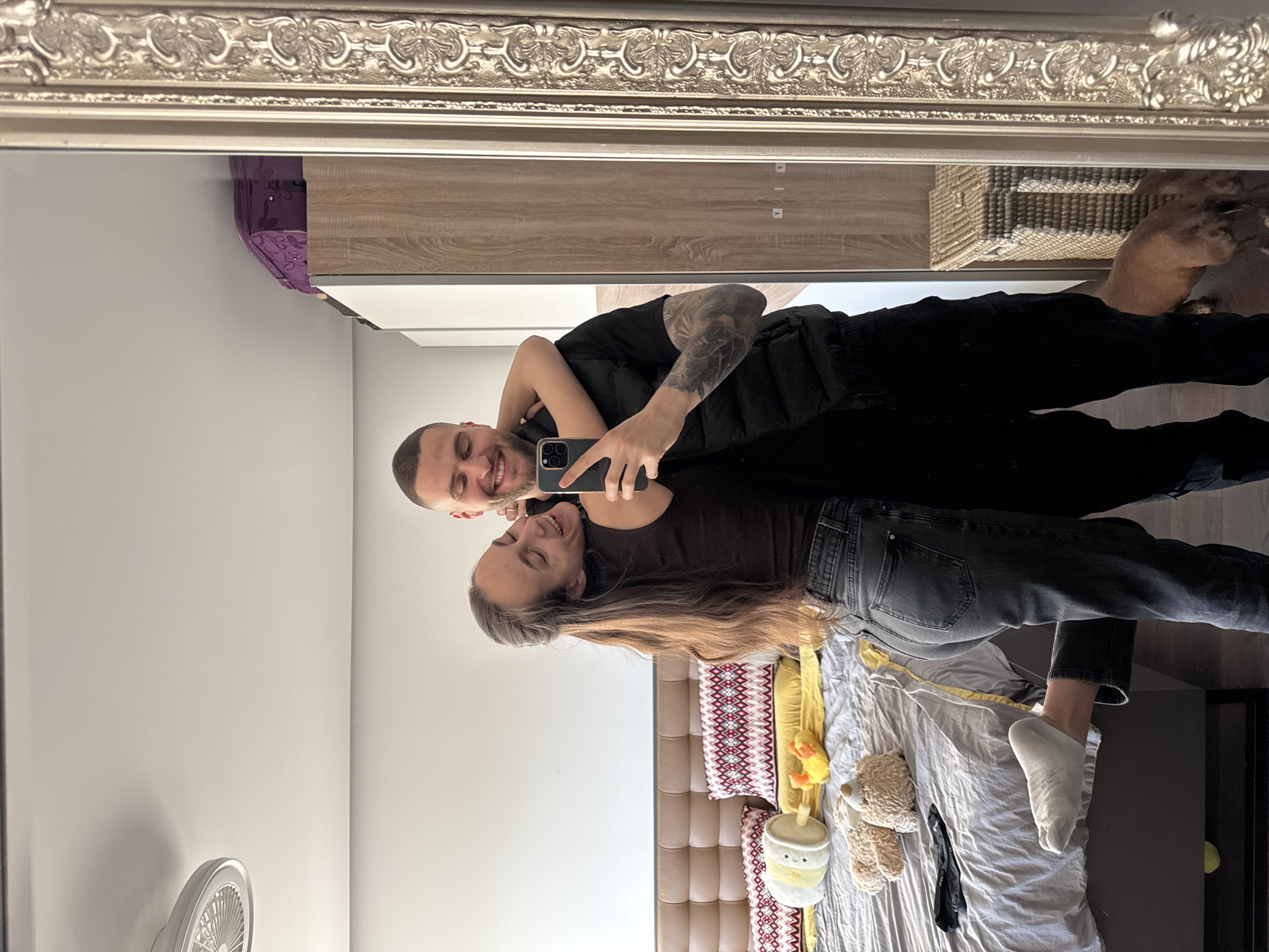 This image captures a couple taking a mirror selfie in a bedroom, likely situated in Sofia, Bulgaria. The picture is taken from the perspective of the mirror, which has an ornate, silver-colored frame across the top.
The man, with short dark hair and a beard, is holding a dark-colored smartphone (an iPhone with multiple cameras) in his right hand, angled to capture both individuals. He is wearing a black t-shirt and a dark vest or jacket, showcasing a tattooed left arm. He is smiling widely. The woman, with long brown hair flowing over her shoulder, is leaning into the man, looking towards the camera with a soft smile. She is dressed in a dark t-shirt and dark blue jeans, with one white sock visible on her foot resting on the bed.
The bedroom features a bed with a light brown padded headboard. The bed is covered with a grey and white bedspread, a yellow throw blanket, and multiple pillows, some with a distinctive red and white geometric pattern. Several plush toys are scattered on the bed, including a yellow duck, a brown teddy bear, and a yellow boba tea-shaped character. Other visible items include a white wall-mounted fan on the left, a wooden cabinet or dresser in the background, and a woven basket atop it. The couple's joyful expressions convey happiness and affection.