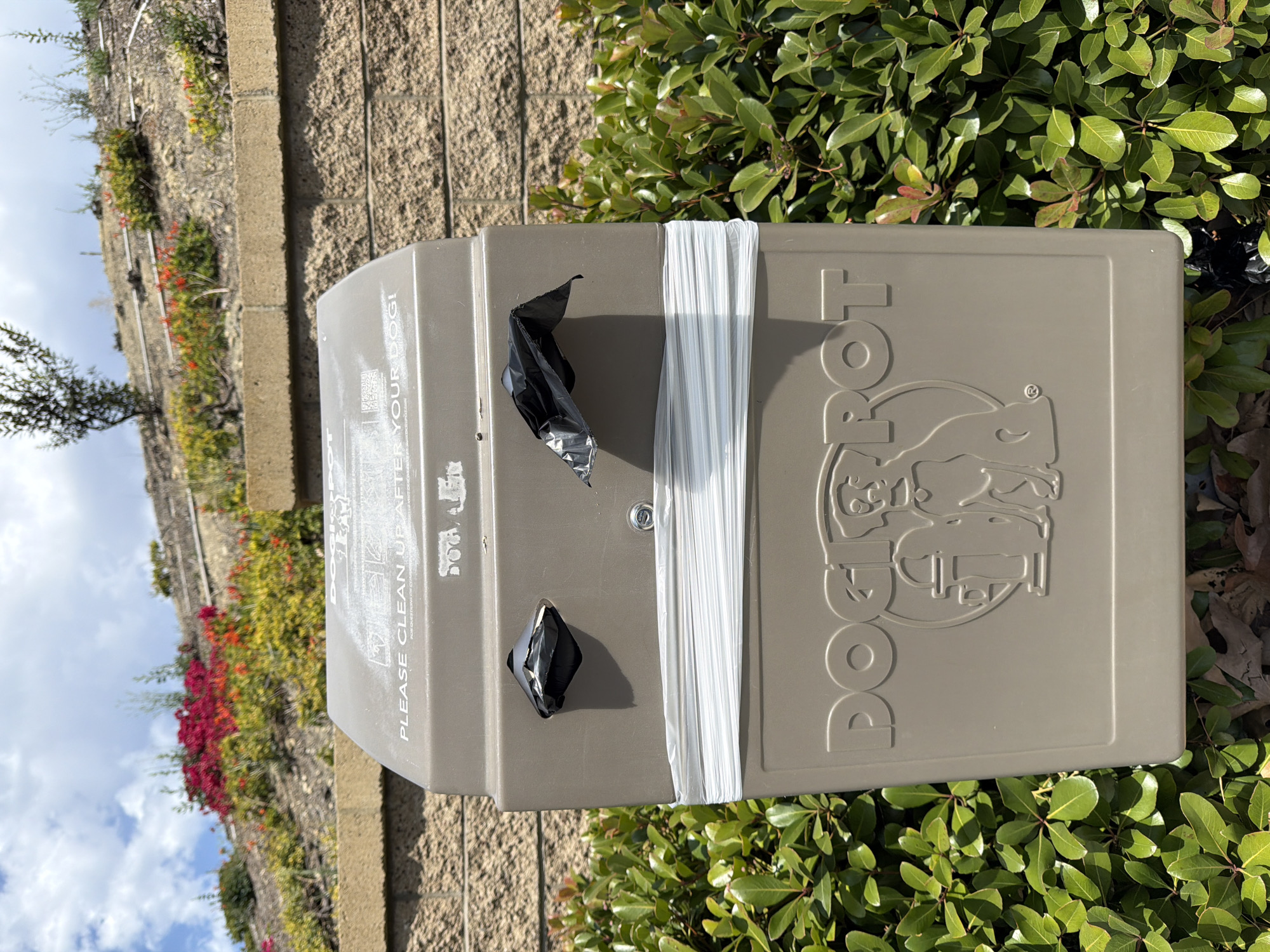 A dog waste station stands in front of a low stone wall, with a slope of flowering bushes and trees behind it, under a cloudy sky. The station is a tan, rectangular box with a domed top. The top of the box has text that reads "PLEASE CLEAN UP AFTER YOUR DOG!". There are two black plastic bags protruding from openings on the front of the station. A large roll of white plastic bags is wrapped around the middle of the station. The front of the station also features the embossed logo "DOGIPOT" with a silhouette of a dog and a trash can. The station is surrounded by green bushes. The background shows a textured brick wall and a raised embankment with landscaping. The location is in Irvine, United States of America.