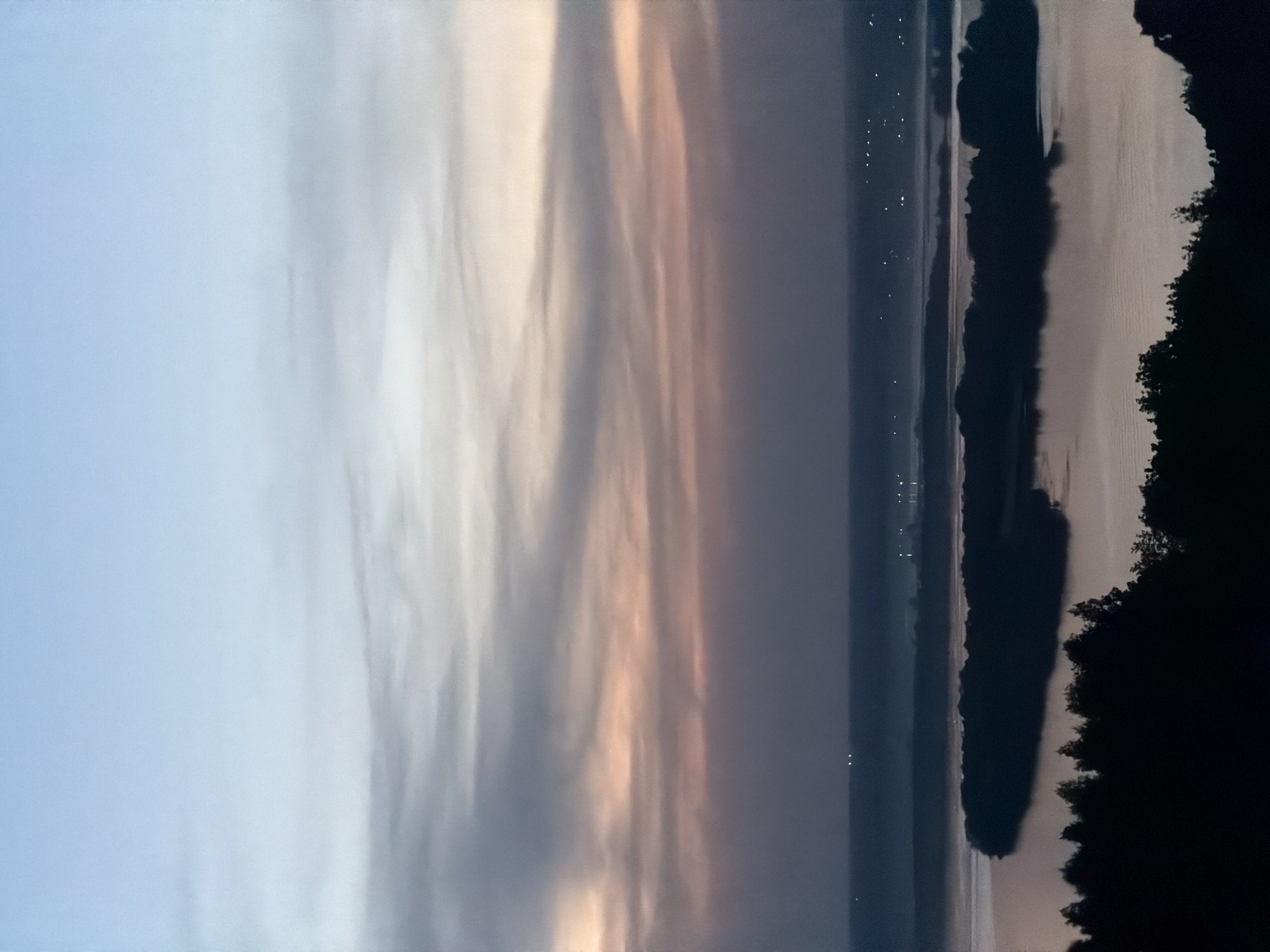 The image shows a body of water, likely a river or lake, at dusk or dawn. The sky is filled with clouds that are illuminated with a mix of grey, white, and peach colors, suggesting the presence of the sun below the horizon. Along the water's edge are dark silhouettes of trees or vegetation. On the opposite shore, faint lights suggest a city or town, potentially Innopolis, Russia. The scene is quiet and still, with a focus on the natural beauty of the landscape.