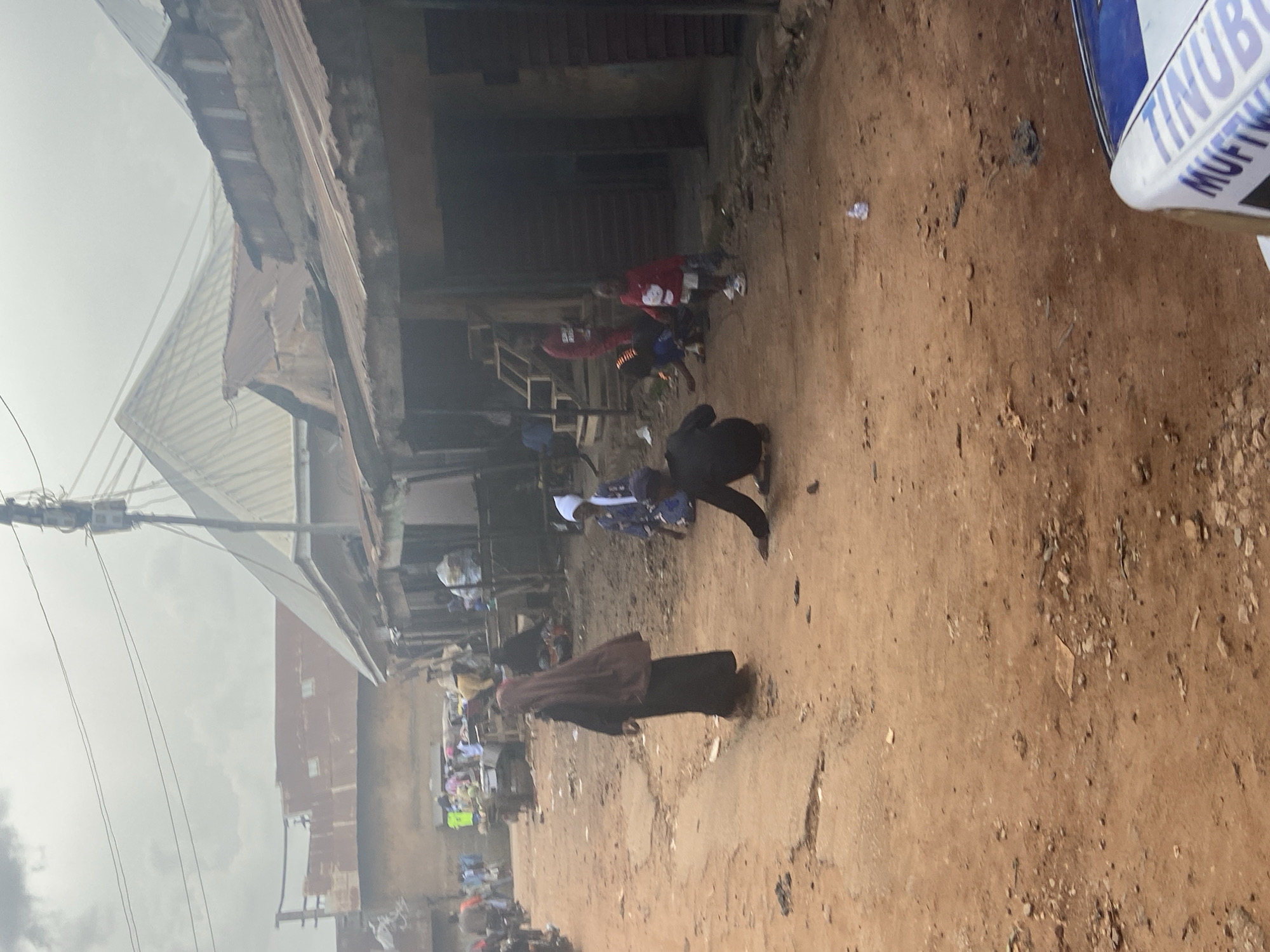 This is a candid, slightly tilted, full shot of a dusty street scene in Bukuru, Nigeria. The foreground is dominated by dry, reddish-brown earth littered with small stones and debris. In the background, a row of buildings with corrugated metal roofs and wooden structures line the street. The buildings appear weathered and perhaps under construction or in disrepair, with exposed beams and rough finishes.
Several people are present. In the mid-ground, a person in a dark garment walks away from the camera. Further back, two individuals in red tops and blue shorts are visible near a wooden staircase leading into one of the buildings. Other figures are scattered throughout the scene, some appearing to be gathered in groups. The general atmosphere suggests a community setting, possibly a market or a residential area.
The sky above is overcast, with visible clouds suggesting diffused light, indicating it is likely daytime, perhaps mid-morning or afternoon. There is no clear indication of a specific event, but the presence of multiple people suggests a normal day in the community. A partial view of a white and blue object with visible text is in the top right corner, but the text is mostly obscured. The visible portion of the text appears to read "TINUBU MUFTW". The overall impression is of a bustling, perhaps impoverished, urban environment.