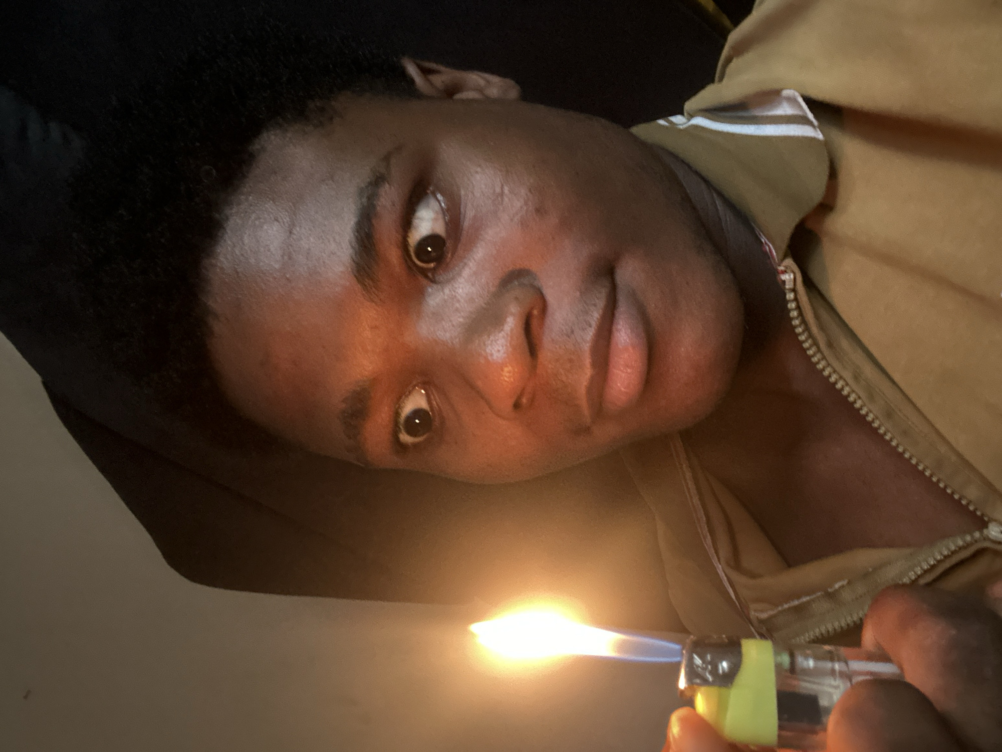 In Ihiagwa, Nigeria, a young Black male is holding a lit lighter in his left hand, positioned to the left of the frame. The lighter is a clear plastic and yellow disposable model with a visible flame. The male's face is angled towards the right, with his eyes wide open and looking off to the right. His facial expression suggests surprise or shock. He is wearing a tan-colored zip-up jacket with white and blue striped trim on the collar. The lighting in the scene is warm and primarily comes from the lighter, casting a glow on his face and the surrounding environment, which appears to be indoors with dark background elements.