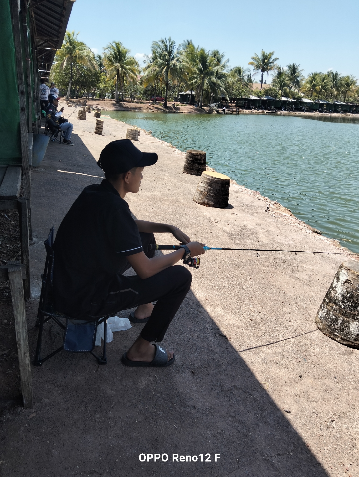 In Cindai Alus, Indonesia, a young male is fishing by the water. He sits on a black folding chair on a concrete walkway, holding a fishing rod with a blue handle. He is wearing a black t-shirt, black pants, and a black baseball cap, with black flip-flops on his feet. The water is calm and green, and palm trees line the opposite bank of the pond. In the background, there are several small fishing huts. The scene is bathed in sunlight, casting a distinct shadow on the walkway.