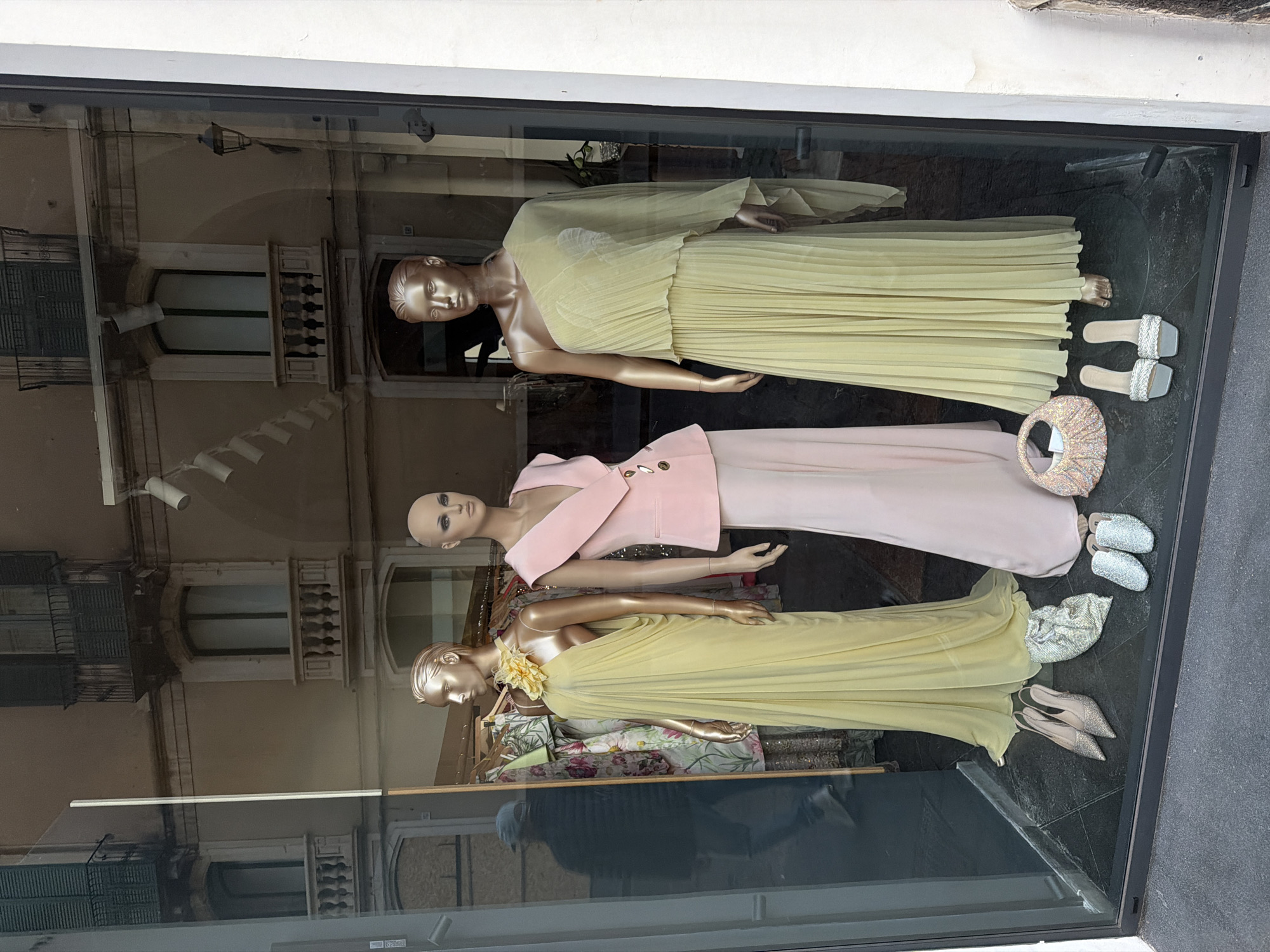 The image shows a window display of a clothing store, likely in Acireale, Italy, based on the reflection of buildings with architectural features typical of the region. Three mannequins are dressed in formal wear. The top mannequin wears a pleated yellow dress, accompanied by a pair of silver and white sandals and a decorative handbag. The middle mannequin is dressed in a pale pink dress with a matching blazer featuring decorative buttons. The bottom mannequin is wearing a pale yellow flowing gown with a large yellow flower embellishment, paired with taupe high heels and a beaded bag. The window reflection shows buildings with arched windows, balconies, and street lighting. The display is set against a dark gray floor.