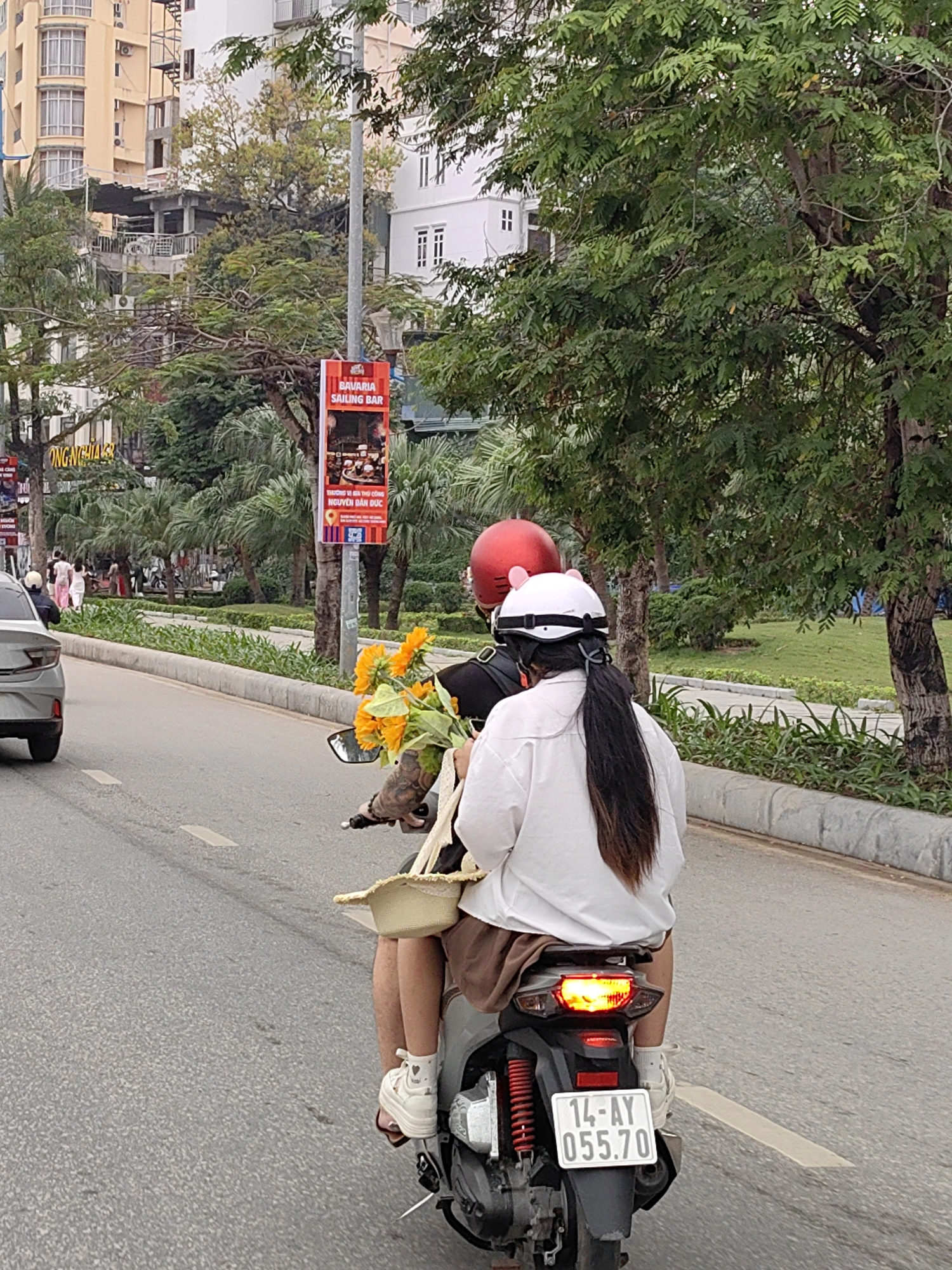 The image captures a daytime urban street scene in Bai Chay Ward, Vietnam. Two people are riding a silver/gray scooter, viewed from behind, traveling along a paved road with white markings. The driver, wearing a red helmet, has a tattooed arm visible on the left side, holding the handlebars. A large bouquet of bright yellow sunflowers and a woven basket are carried on the scooter between the driver and the handlebars.
The passenger, riding pillion, is dressed in a white long-sleeved shirt, brown skirt or shorts, and white platform sneakers with heart-shaped details. She wears a white helmet adorned with pink cat ears, and her long dark hair is tied in a ponytail. The scooter's license plate, clearly visible at the rear, reads "14-AY 055.70."
To the left of the scooter, a gray car is partially visible. The road is bordered by a concrete curb and a narrow strip of grass, beyond which lies a wide sidewalk with several pedestrians. Lush green trees, including palm trees, line the sidewalk and a central median.
In the background, multi-story buildings, some with balconies, are visible through the foliage. A tall street light pole features two red advertising banners. The top banner prominently displays "BAVARIA SAILING BAR." The lower banner contains Vietnamese text: "THƯƠNG VỊ ĐỀU THỦ CÔNG" (Handicraft Flavor) and "NGUYÊN DÂN ĐỨC" (Original German). Further down the street, another yellow sign shows the text "ONG NGHIA." The lighting suggests an overcast day or late afternoon, creating a soft, even illumination.