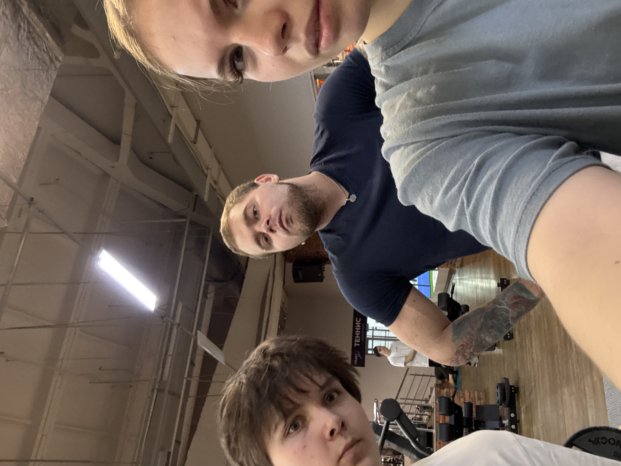 A selfie-style, slightly tilted, indoor shot featuring three people at a gym in Chaikovsky, Russia. A fair-skinned woman with blonde hair is in the upper left of the frame. Below and slightly to the right of her is a fair-skinned man with a beard and a dark-colored shirt, displaying an arm tattoo. Further down in the foreground is a person with short, dark hair and a white shirt. The gym interior includes exercise equipment, a bright ceiling light, a sign in Russian, and another person in the background.