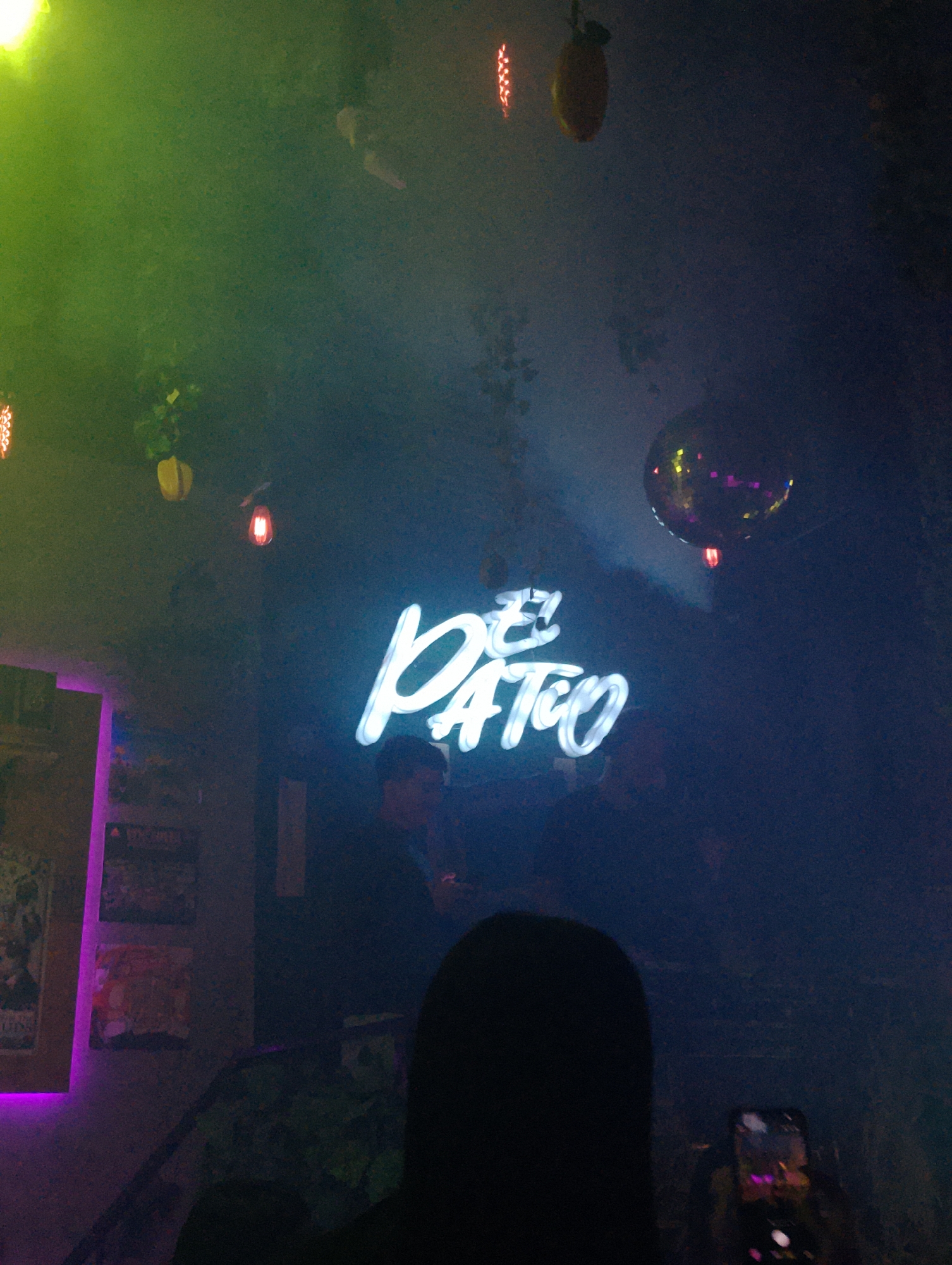 The image depicts a dimly lit interior space, likely a bar or club, with a neon sign that reads "El Patio" in a stylized script. The ambient lighting is colorful, with green and purple hues visible. People are present in the background, though their features are indistinct. A disco ball hangs from the ceiling. In the foreground, a person's head is silhouetted, and another person is holding a phone to record. Decorations include hanging plants and what appear to be lemons. Posters or artwork are mounted on a wall in the background. The location is inferred to be Porlamar, Venezuela.