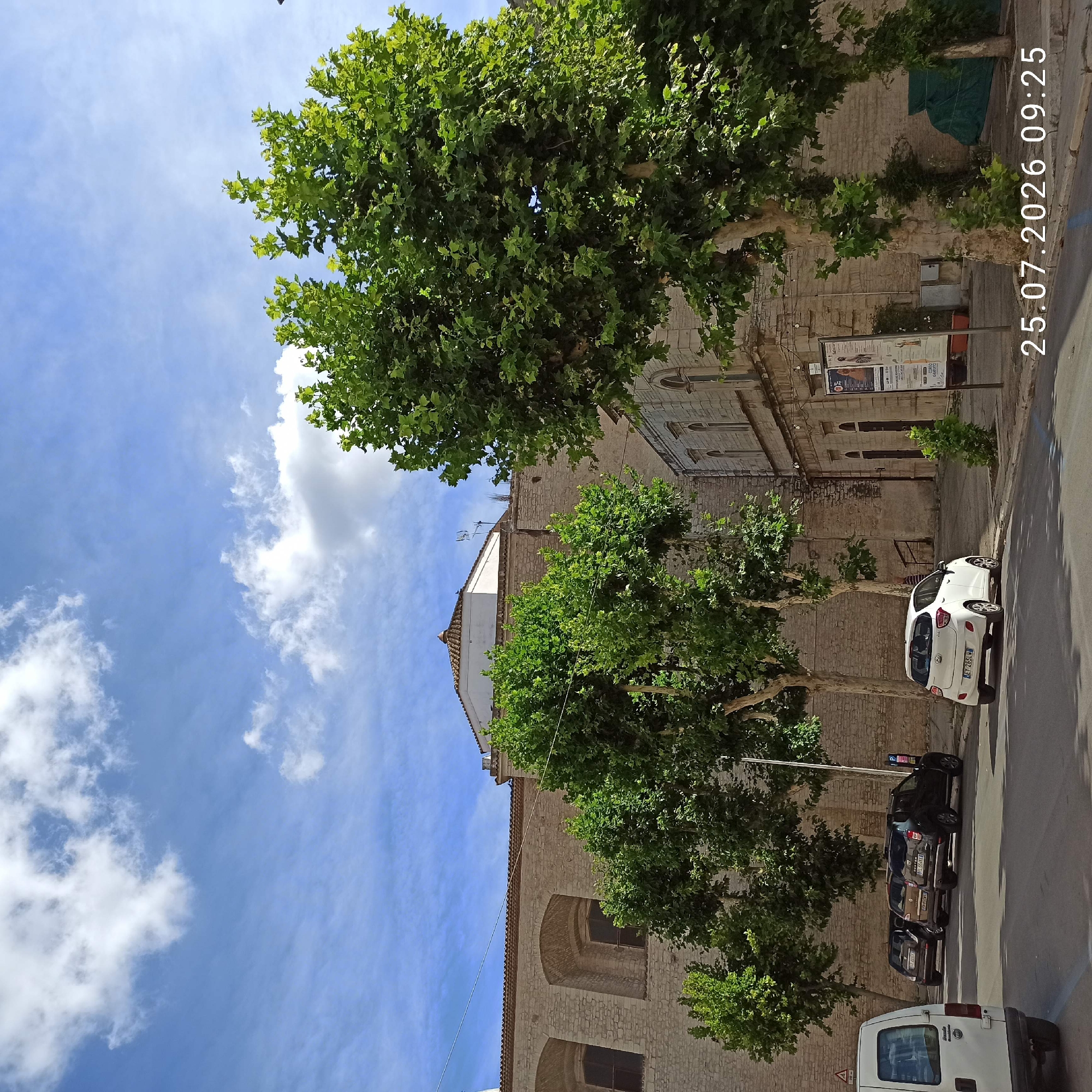 The image shows a street scene in Ruvo di Puglia, Italy, with a large stone building on the left and a smaller building with arched windows on the right. Several trees with lush green foliage are prominent in the foreground and middle ground. There are several parked cars on the street, including a white hatchback, a dark SUV, and other vehicles. The sky is blue with scattered white clouds. A timestamp, "25.07.2026 09:25," is overlaid on the bottom right of the image. The overall impression is a sunny day in a historic Italian town.