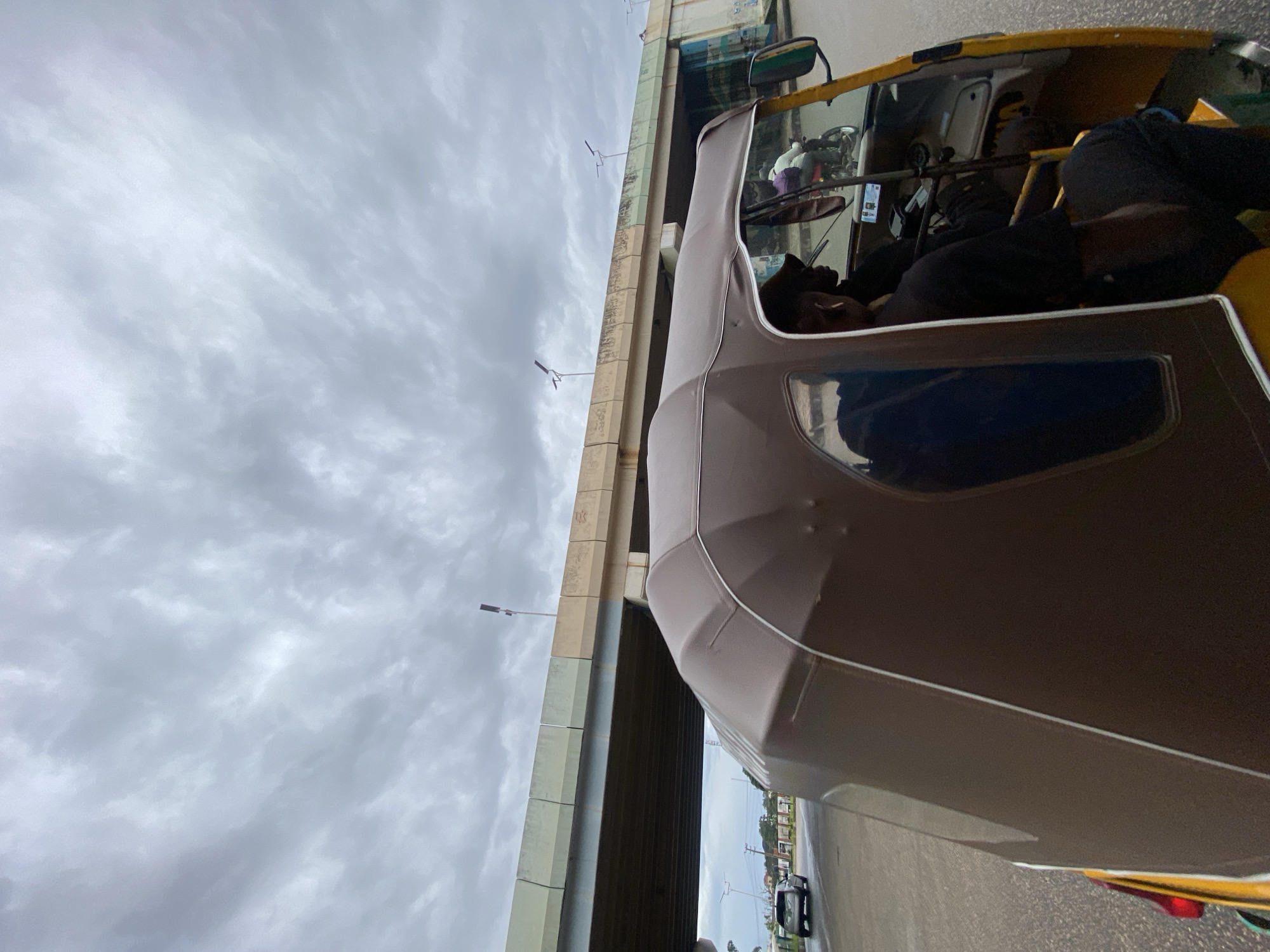 The image shows a view from inside a motorized rickshaw, also known as an "okada" in Nigeria, as it travels on a road. The rickshaw has a brown canopy and yellow sides. Inside, a person wearing a dark t-shirt and a baseball cap is visible, apparently driving the vehicle. Another person is partially visible in the passenger seat. In the background, a large concrete overpass is visible, with streetlights mounted on its sides. The sky is overcast with grey clouds. A car and a motorcycle are also seen on the road. The overall scene suggests an urban environment in Jos, Nigeria, with typical road infrastructure and transportation.