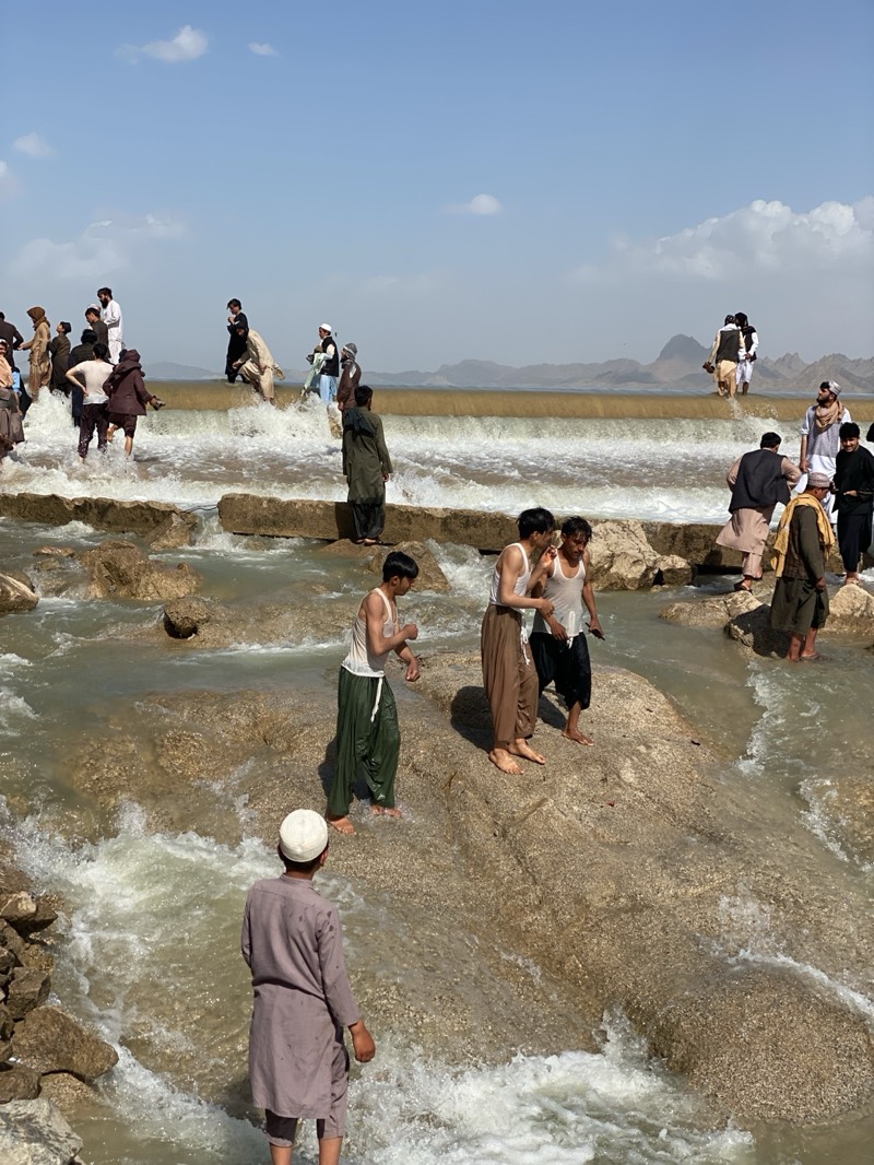 A large group of men are near a body of water on a sunny day in Kim, Afghanistan. Some men are wading in the water, while others are standing on rocks or on a man-made dam-like structure. Most of the men are wearing traditional Afghan clothing, including loose pants and tunics. Some men are wearing white caps or turbans. The water is flowing rapidly and is churning in some areas. In the background, there are low-lying mountains. The sky is blue with some clouds.