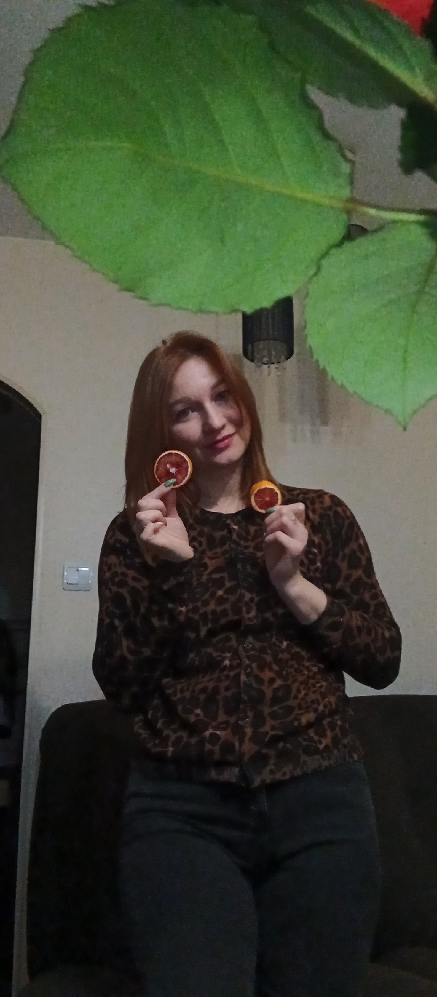 The image shows a woman indoors. She has red hair and is wearing a brown and black patterned cardigan and dark jeans. She is holding two slices of blood orange up in front of her face. Behind her, there is a dark couch and a white wall with a light switch. Large green leaves are visible in the upper portion of the frame. A black lampshade is hanging from the ceiling. The location is Unknown, Unknown.