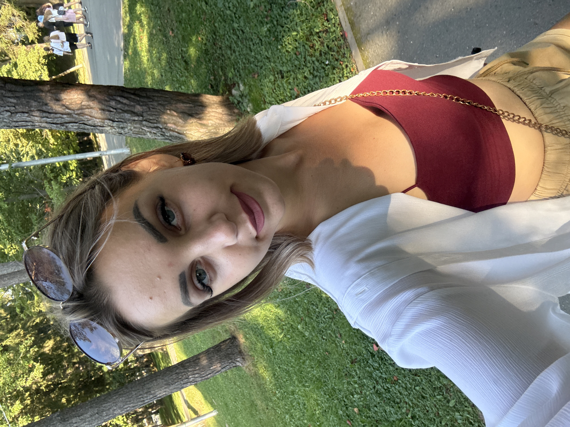 The image shows a young woman with blonde hair, wearing sunglasses, a white open shirt over a burgundy top, and light-colored shorts. She is holding a phone and taking a selfie in a park. The background shows trees, green grass, and a paved path where several people are walking in the distance. The lighting suggests it is daytime, possibly late afternoon due to the shadows. The location is identified as Yuzhno-Sakhalinsk, Russia.